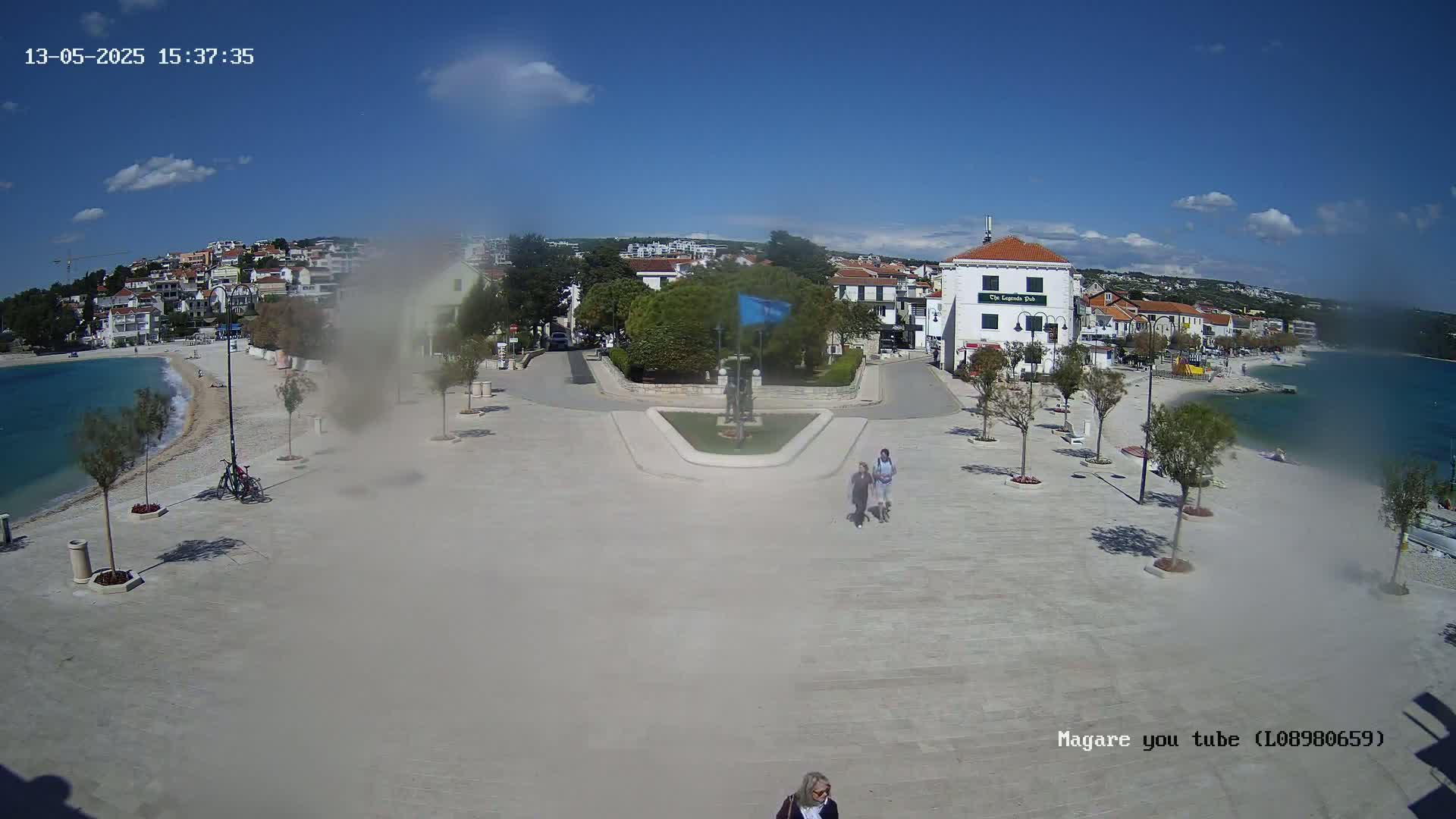 A seaside town square with a small beach, buildings, and a few people walking on a sunny day.