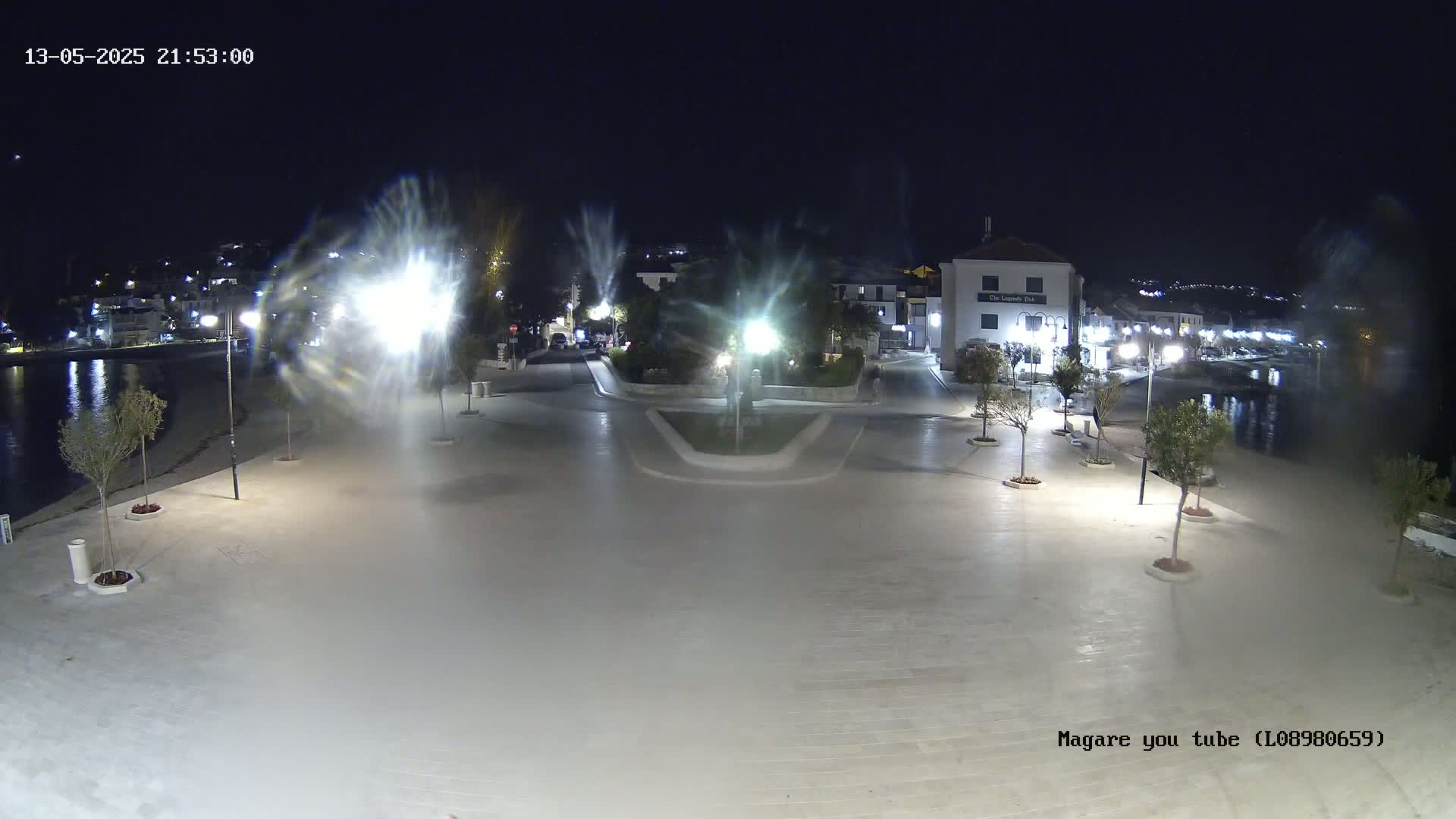 A nighttime, brightly lit town square near a body of water, with small trees planted along the paved area.