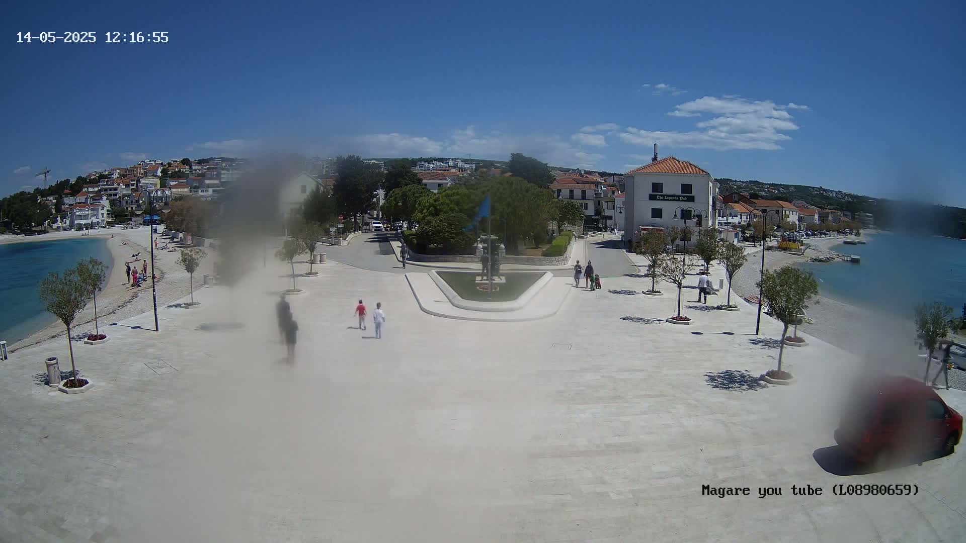 A hazy, sunny day overlooks a town square by the sea, where people stroll near a small beach and a central fountain.