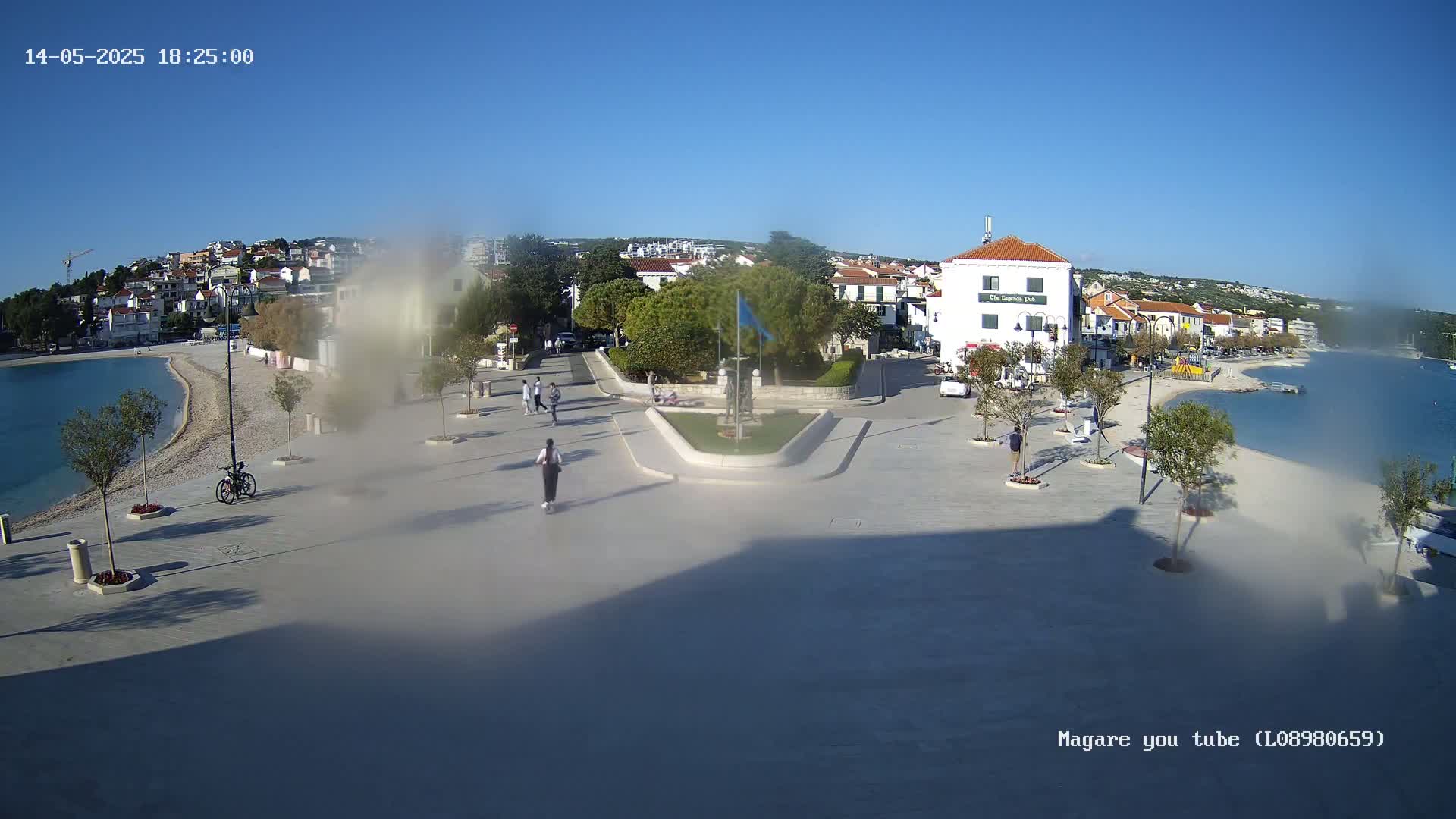 A paved plaza overlooks a calm bay, with a few people strolling near white buildings under a clear blue sky.