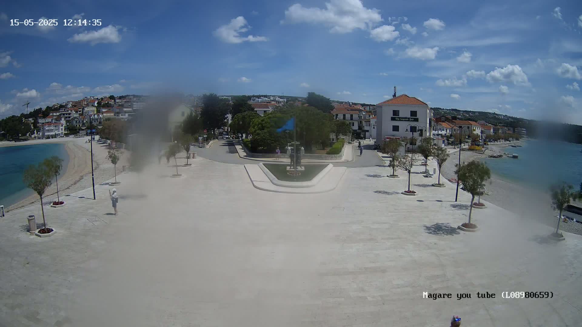 A paved town square overlooks a calm bay on a sunny day, with buildings and trees lining the waterfront.