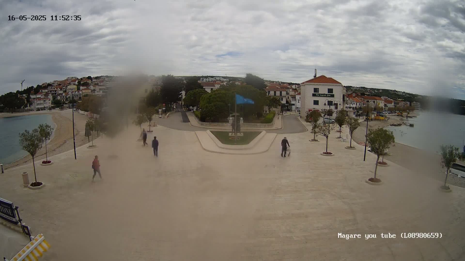 A hazy, overcast day reveals a seaside town square with a few pedestrians, a small beach, and buildings lining a calm bay.