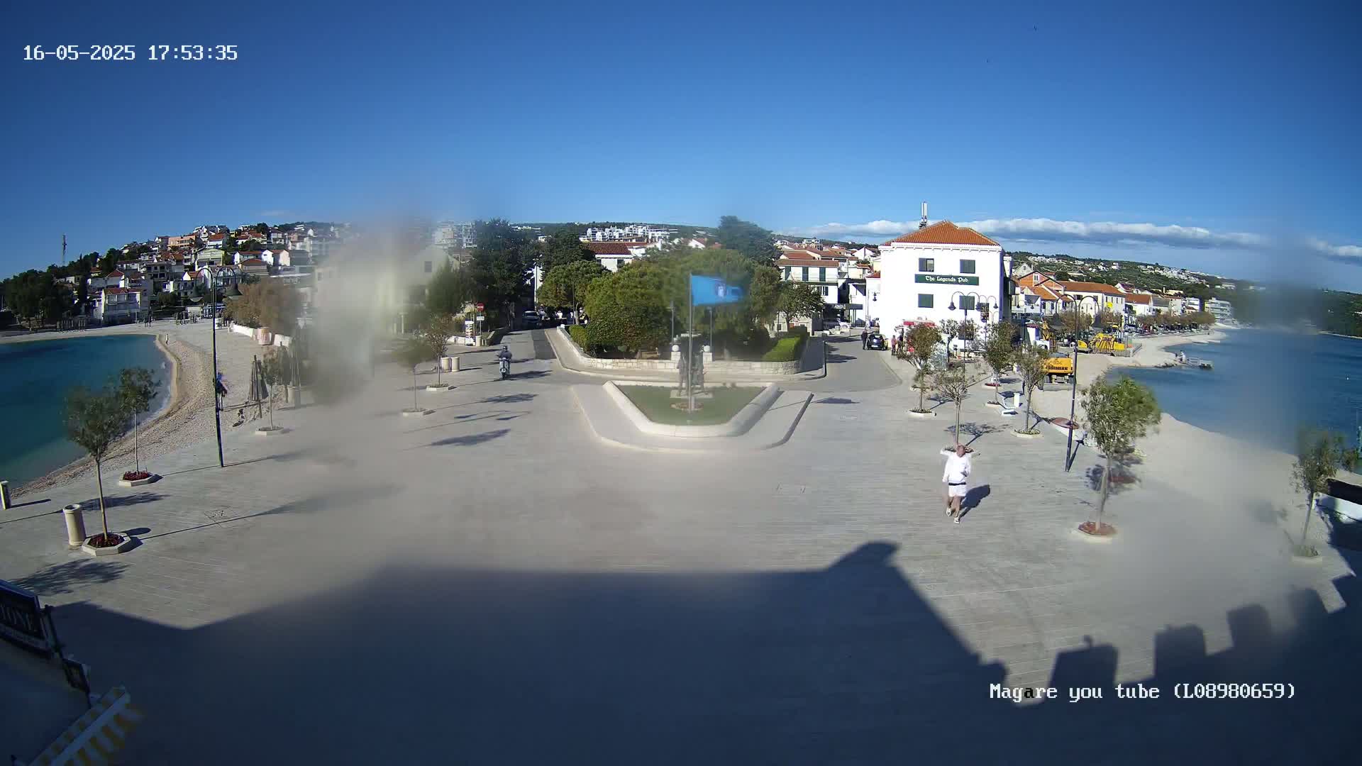 A seaside town square with a small monument in the center, a few people walking, and buildings along a clear, sunny day.