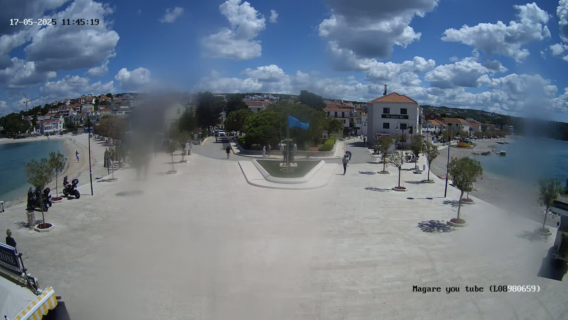 A mostly sunny day shows a town square overlooking a calm bay, with several people walking around and a few vehicles parked.