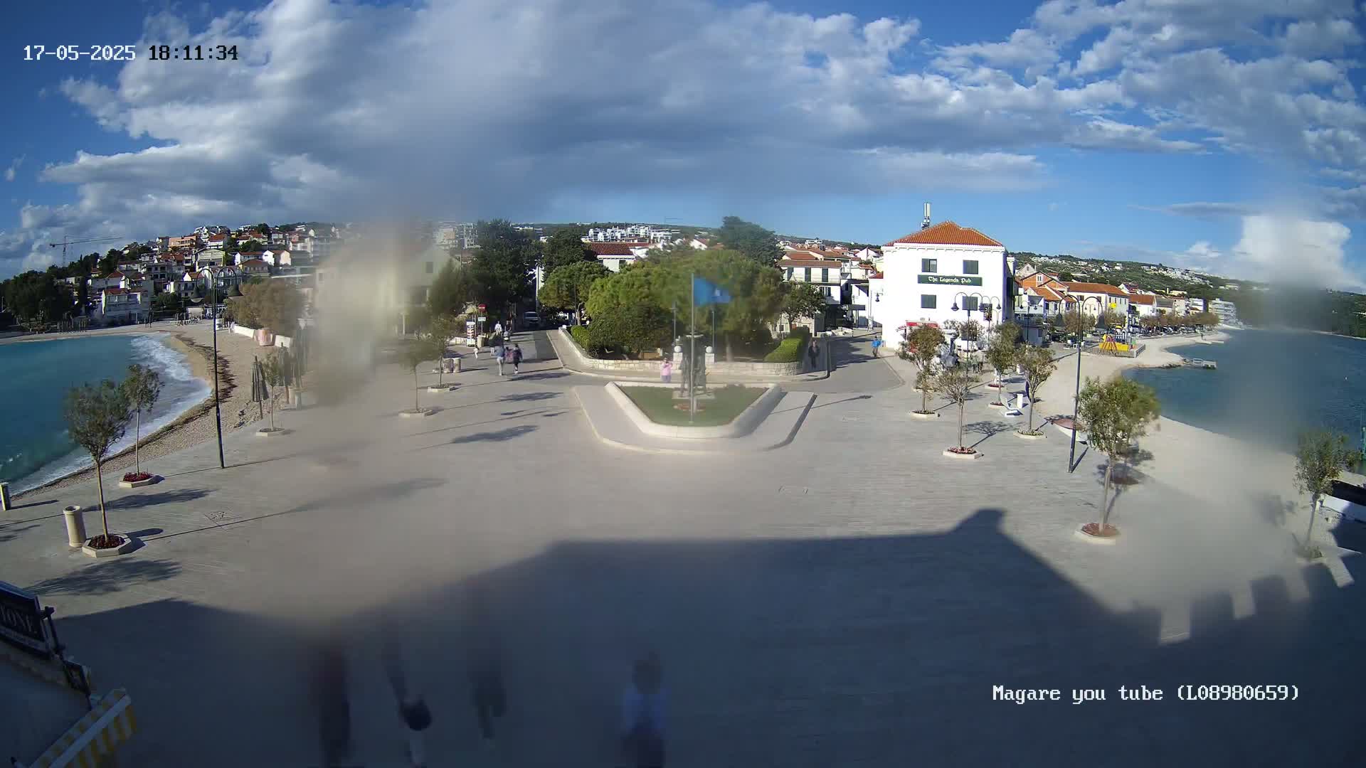 A mostly sunny seaside town square with a small beach and calm water, featuring a central monument and a few people walking around.