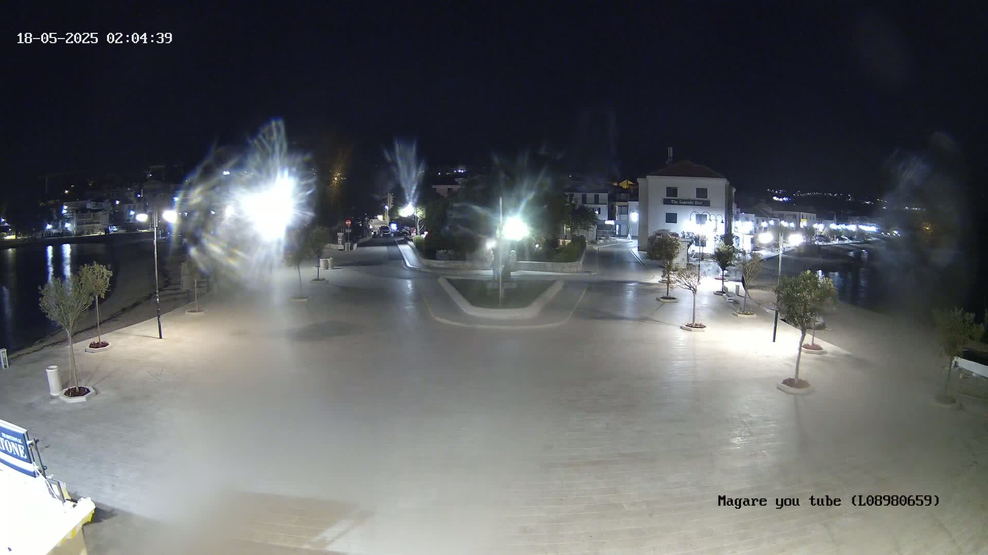 A nighttime, brightly lit plaza overlooks a calm body of water, with buildings and trees visible in the background.