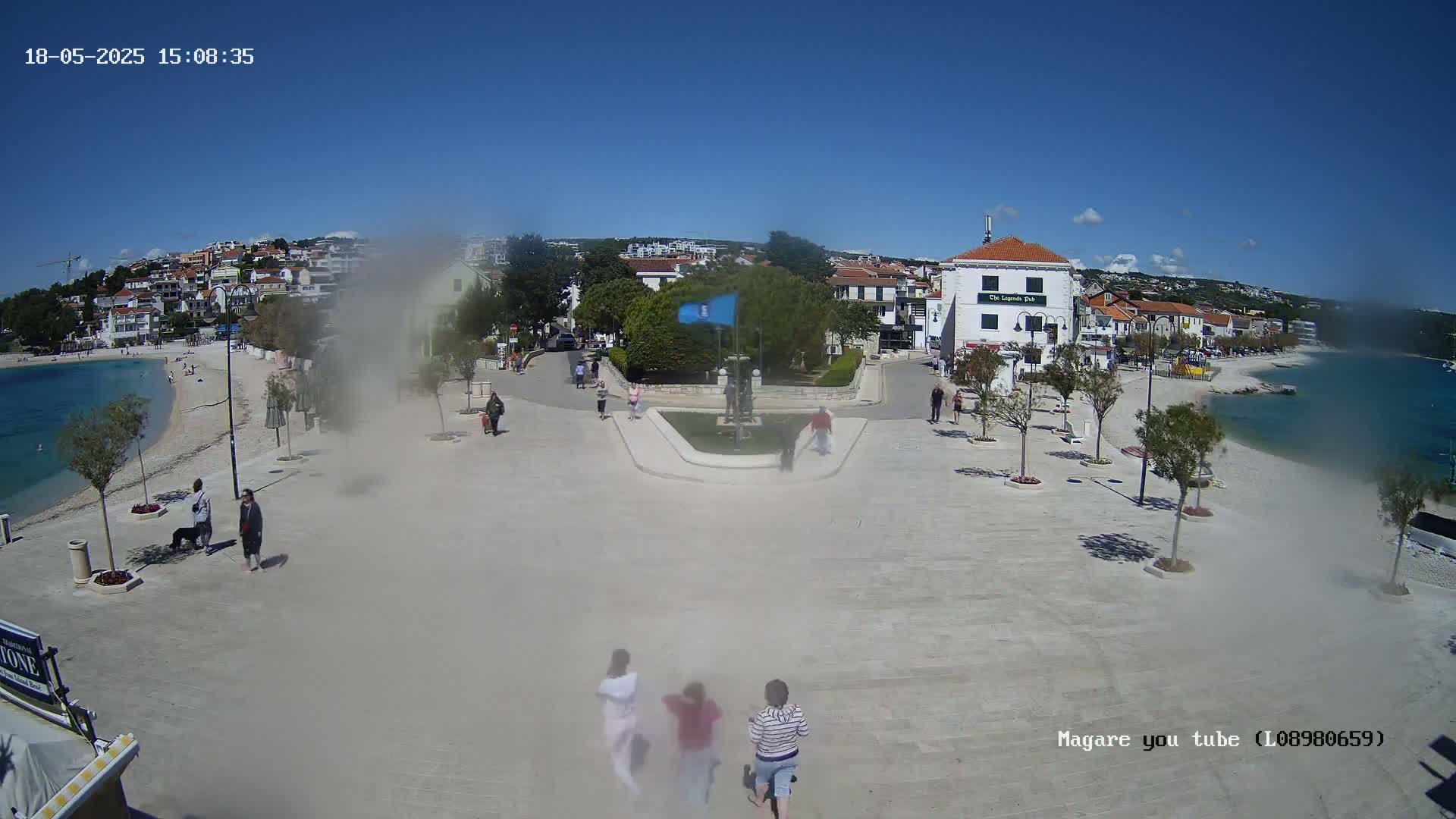 A paved plaza near a calm, blue body of water is filled with people under a clear, sunny sky; buildings and trees surround the plaza.