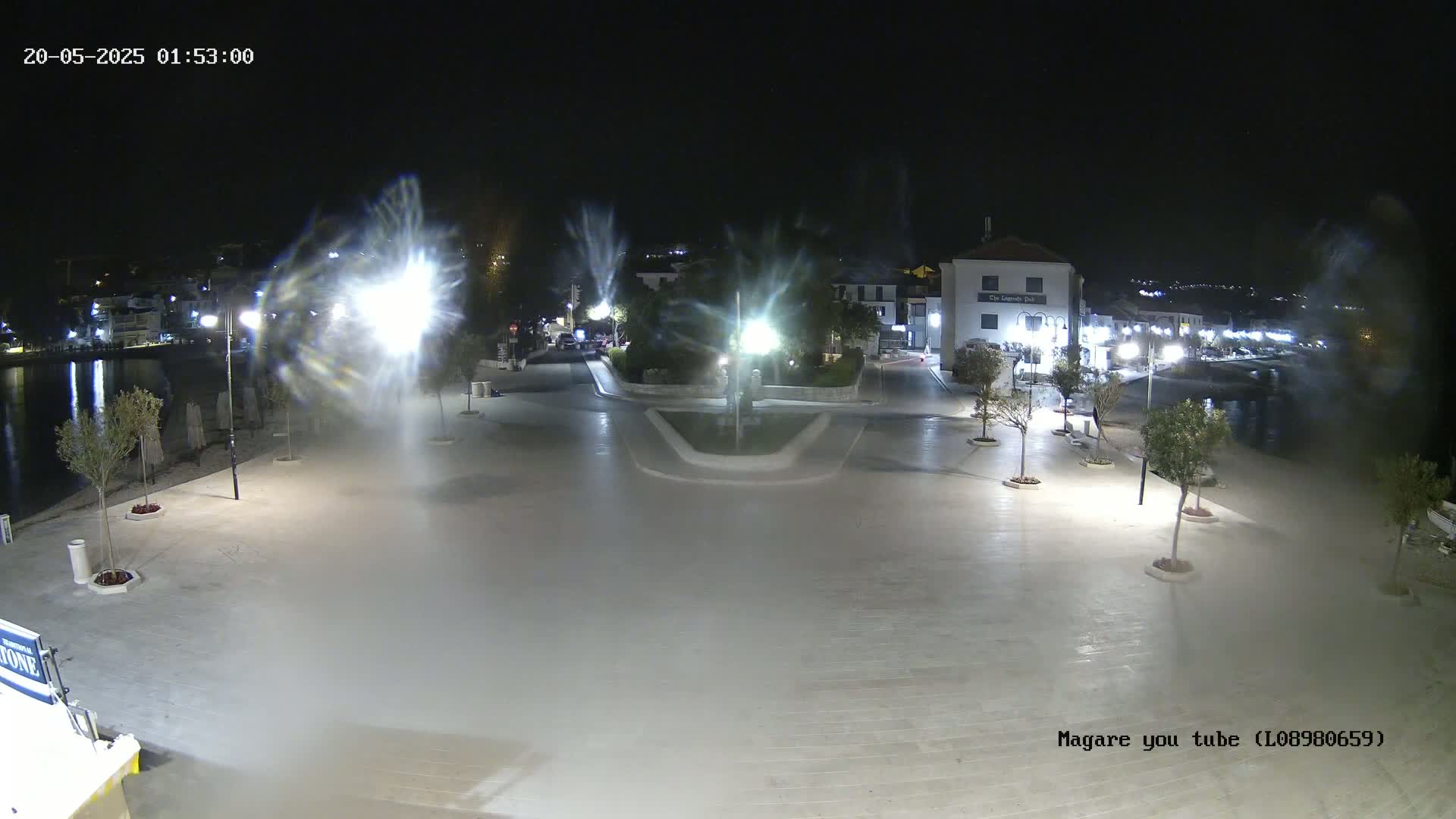 A nighttime, brightly lit plaza near a body of water shows several small trees in planters and buildings in the background.