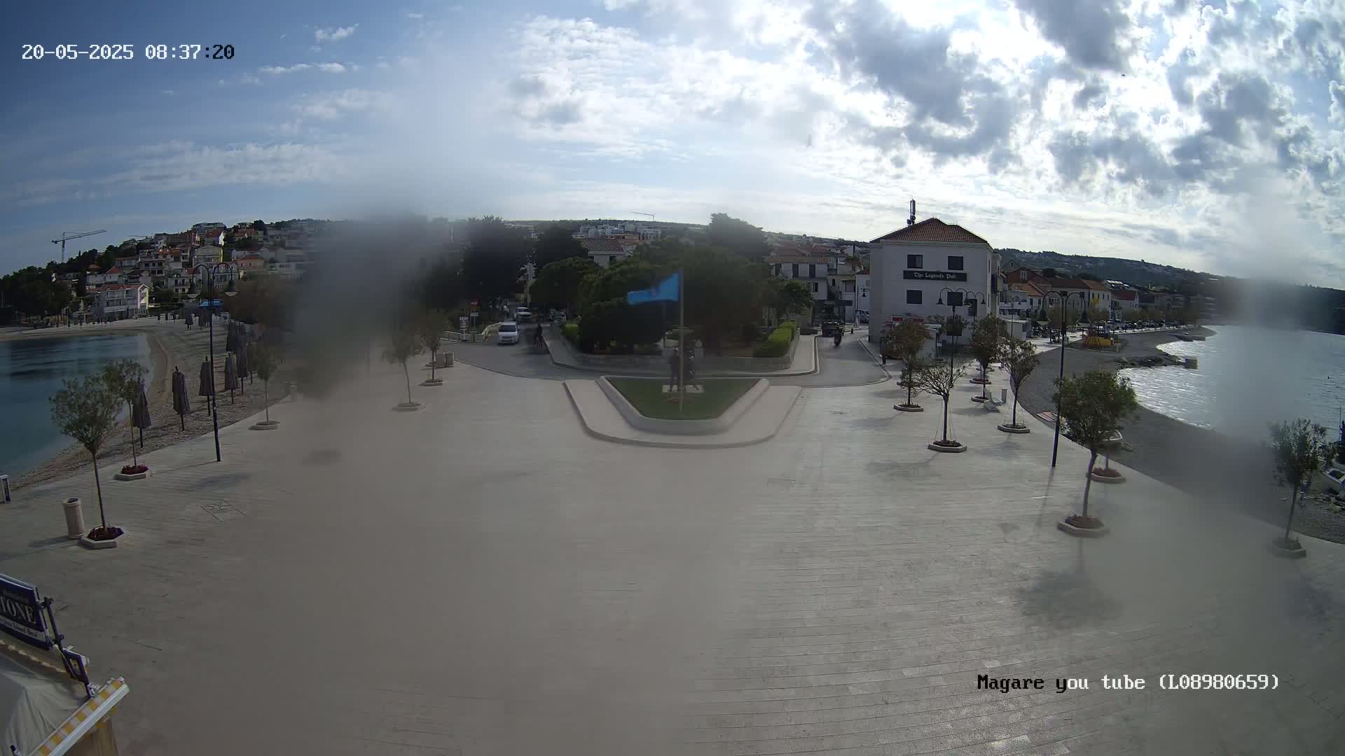 A hazy, partly cloudy day reveals a seaside town square with a calm body of water, a beach, and buildings in the background.