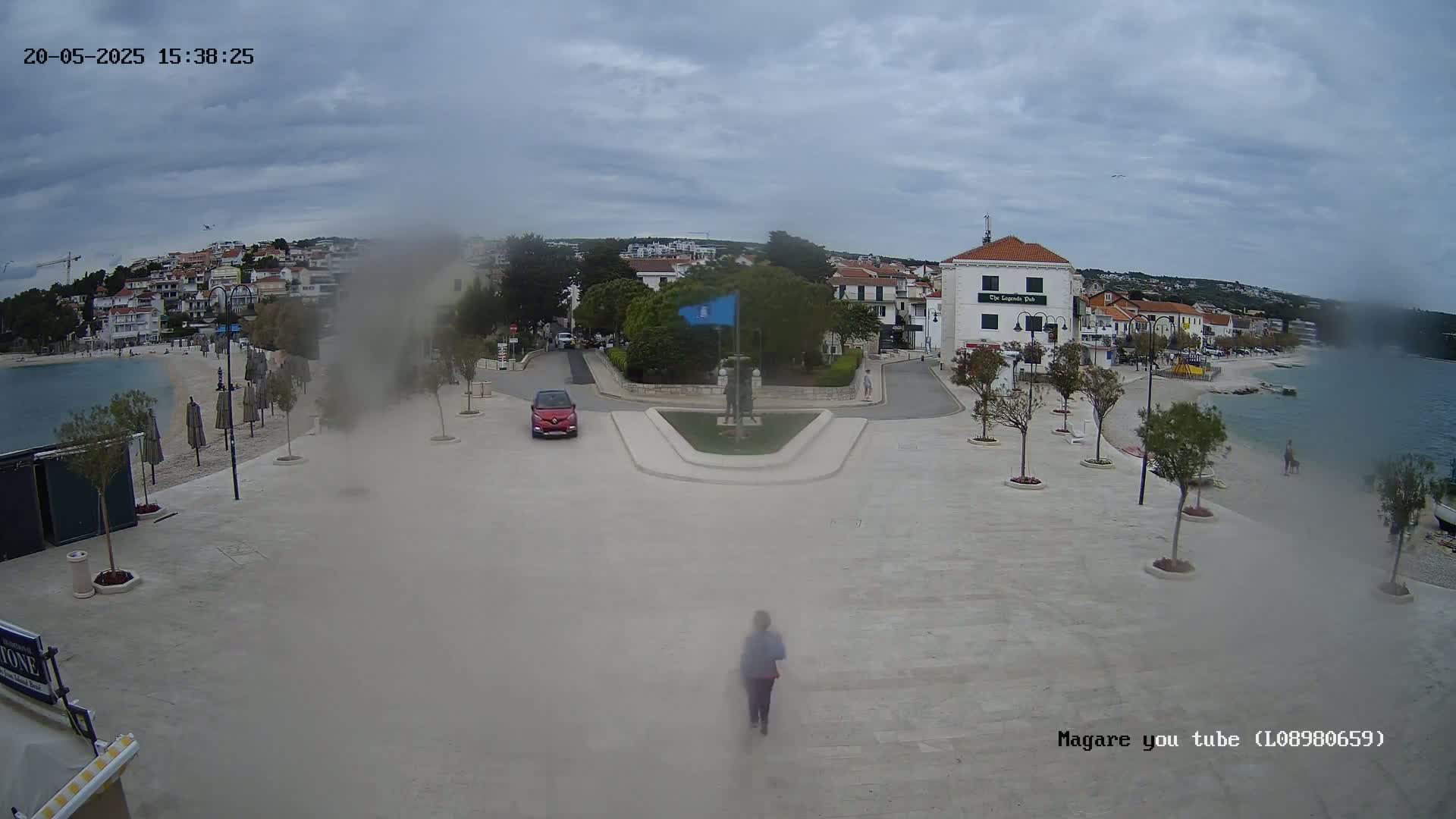 A hazy overcast day reveals a seaside town square with a small red car, a person walking, and a monument in the center, bordered by buildings, trees, and a beach.