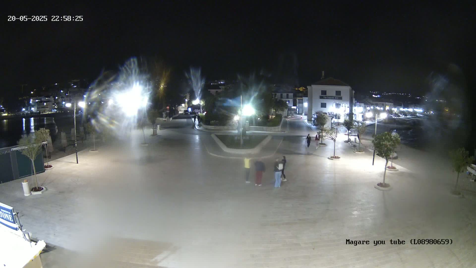 A nighttime, hazy scene shows a paved waterfront plaza with a few people, trees in planters, and buildings, illuminated by bright lights.