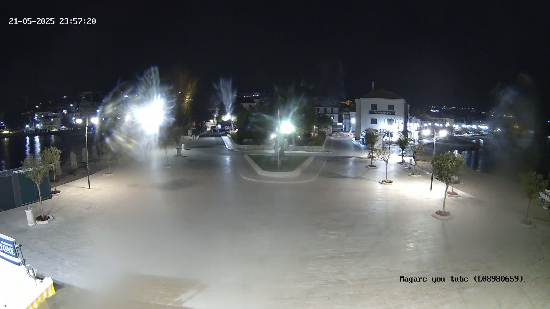 A nighttime, brightly lit plaza near a body of water, with several trees and buildings visible in the background.