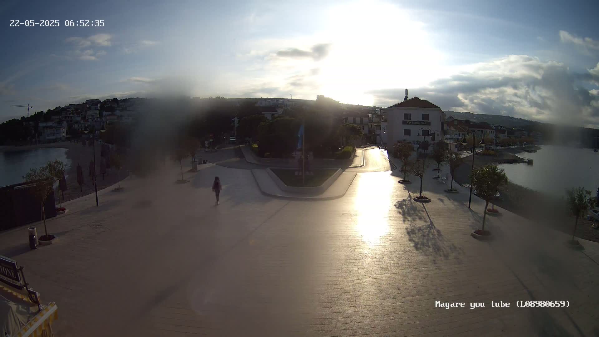A sunlit seaside town square, with a person walking on a paved area near the water, under a partly cloudy sky.