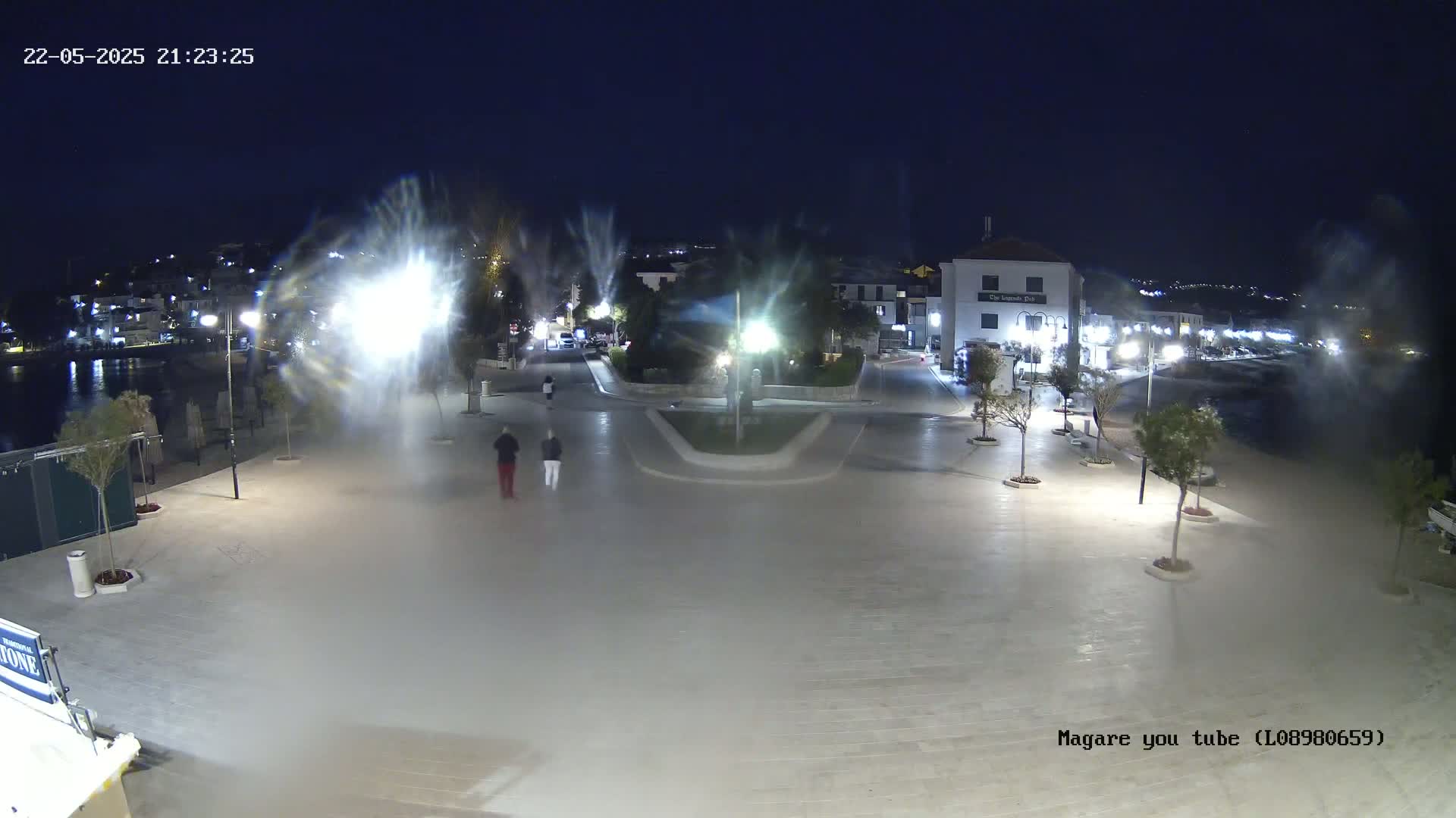 A nighttime view of an illuminated town square near a waterfront, with some water spray visible and two pedestrians walking.