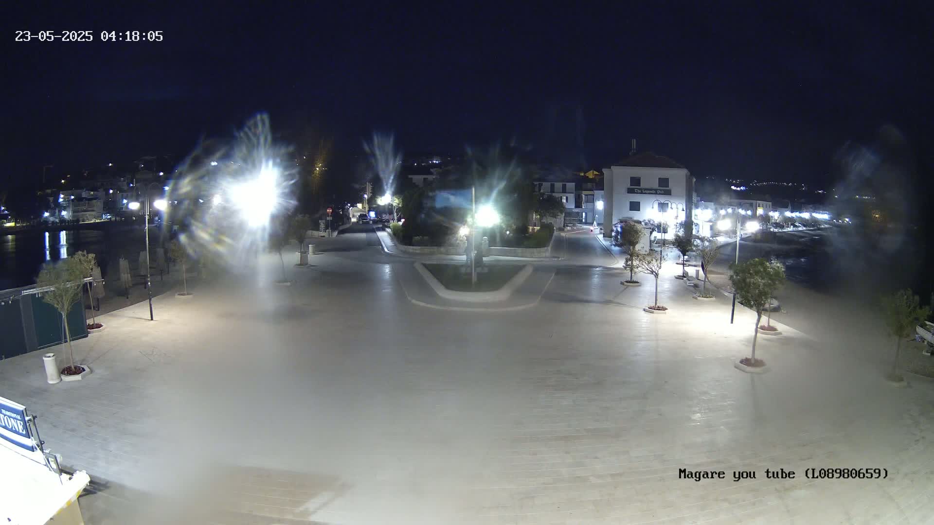 A nighttime view of a town square near a body of water, illuminated by streetlights, with some mist or spray obscuring parts of the scene.