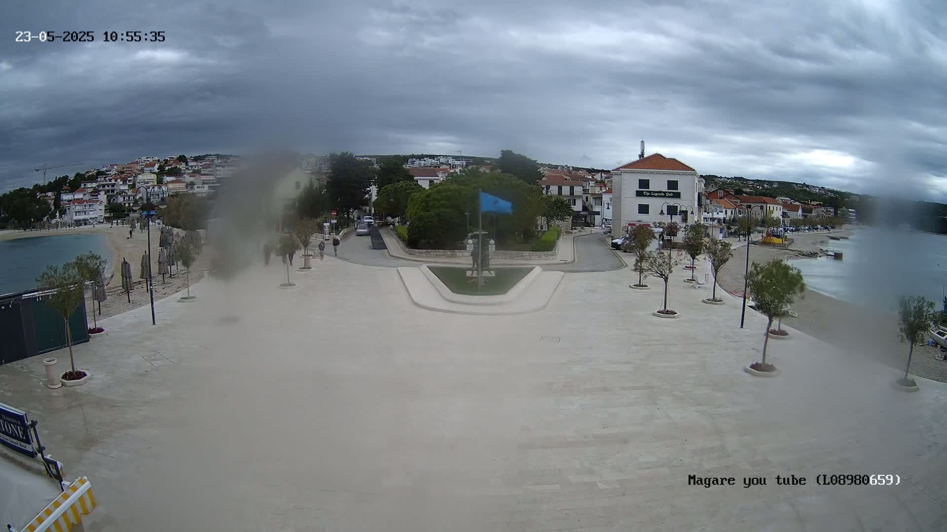 A seaside town square with overcast skies and light rain shows a paved area, small trees in planters, buildings, a small beach, and calm water.