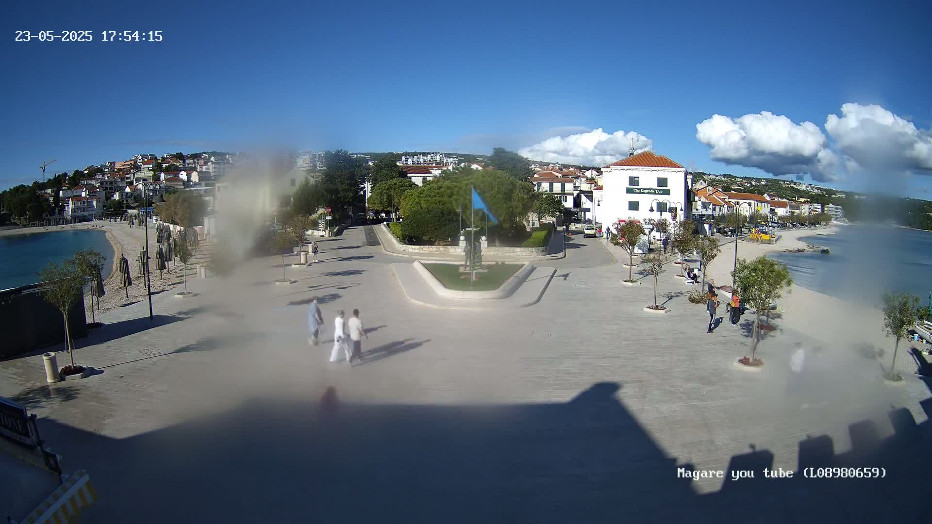 A mostly sunny outdoor scene shows a town square overlooking a calm bay, with several people walking and some buildings visible in the background.