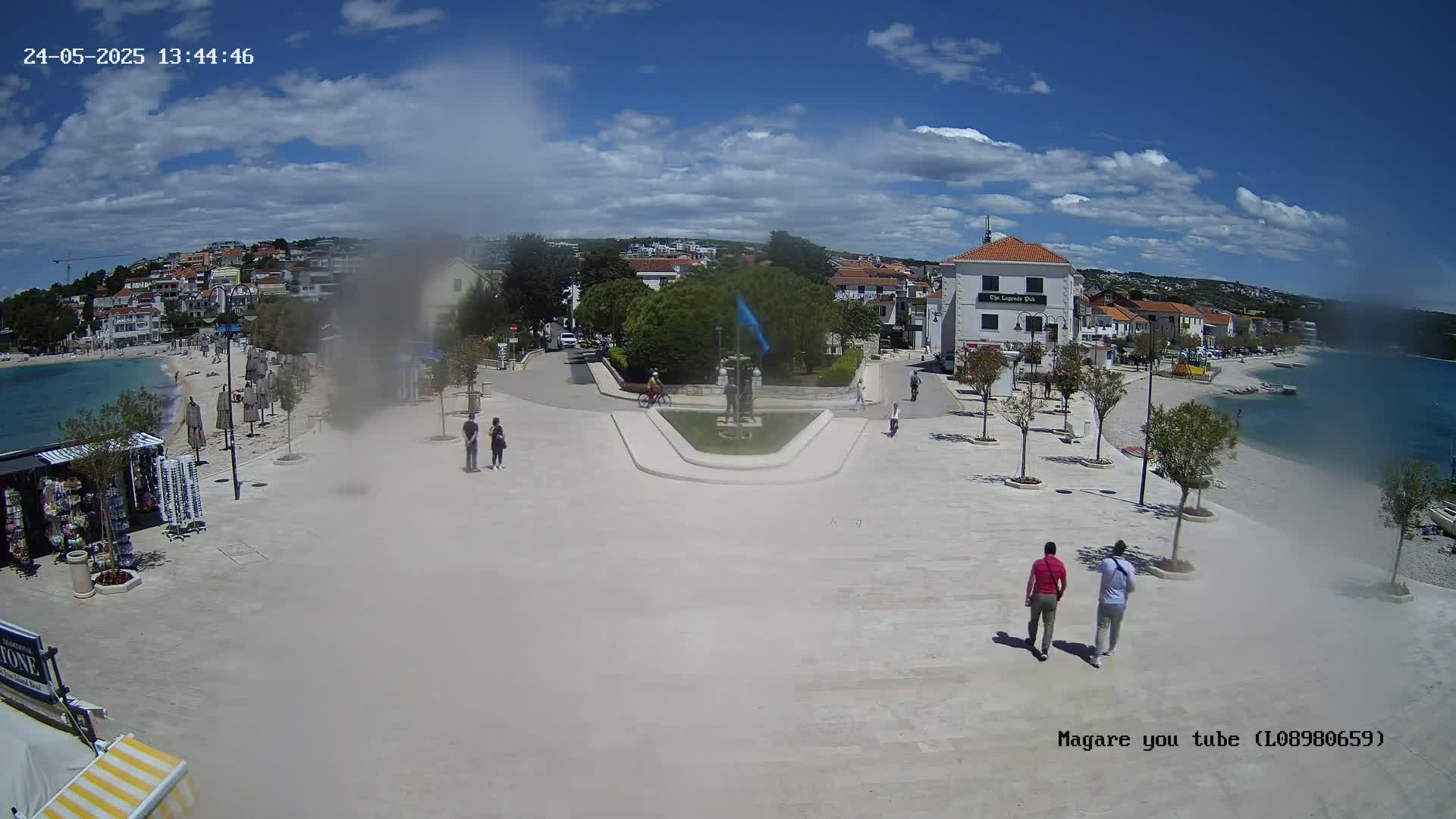 A sunny day overlooking a seaside town square with a few people walking, some small trees, and a calm body of water in the background.