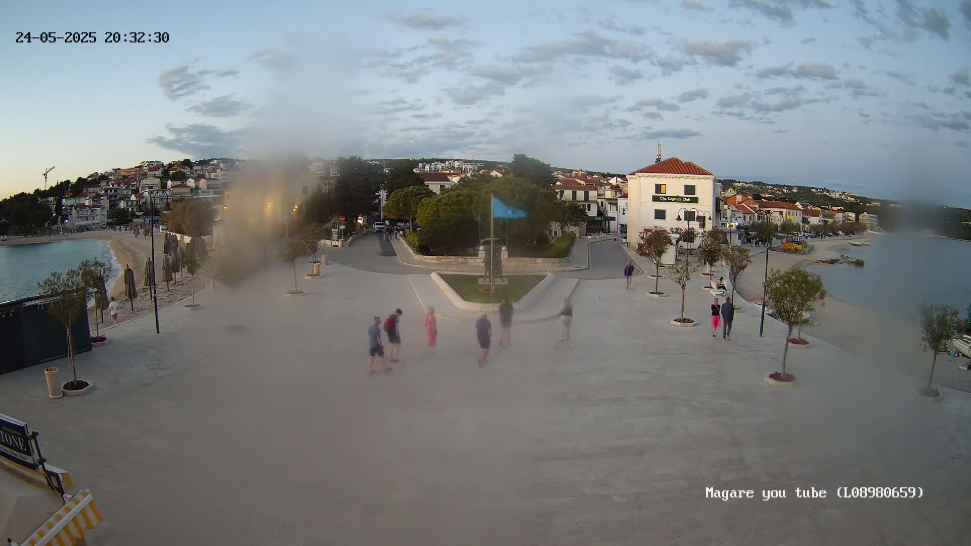 A town square overlooks a calm bay under a partly cloudy evening sky, with several people walking around a central monument.