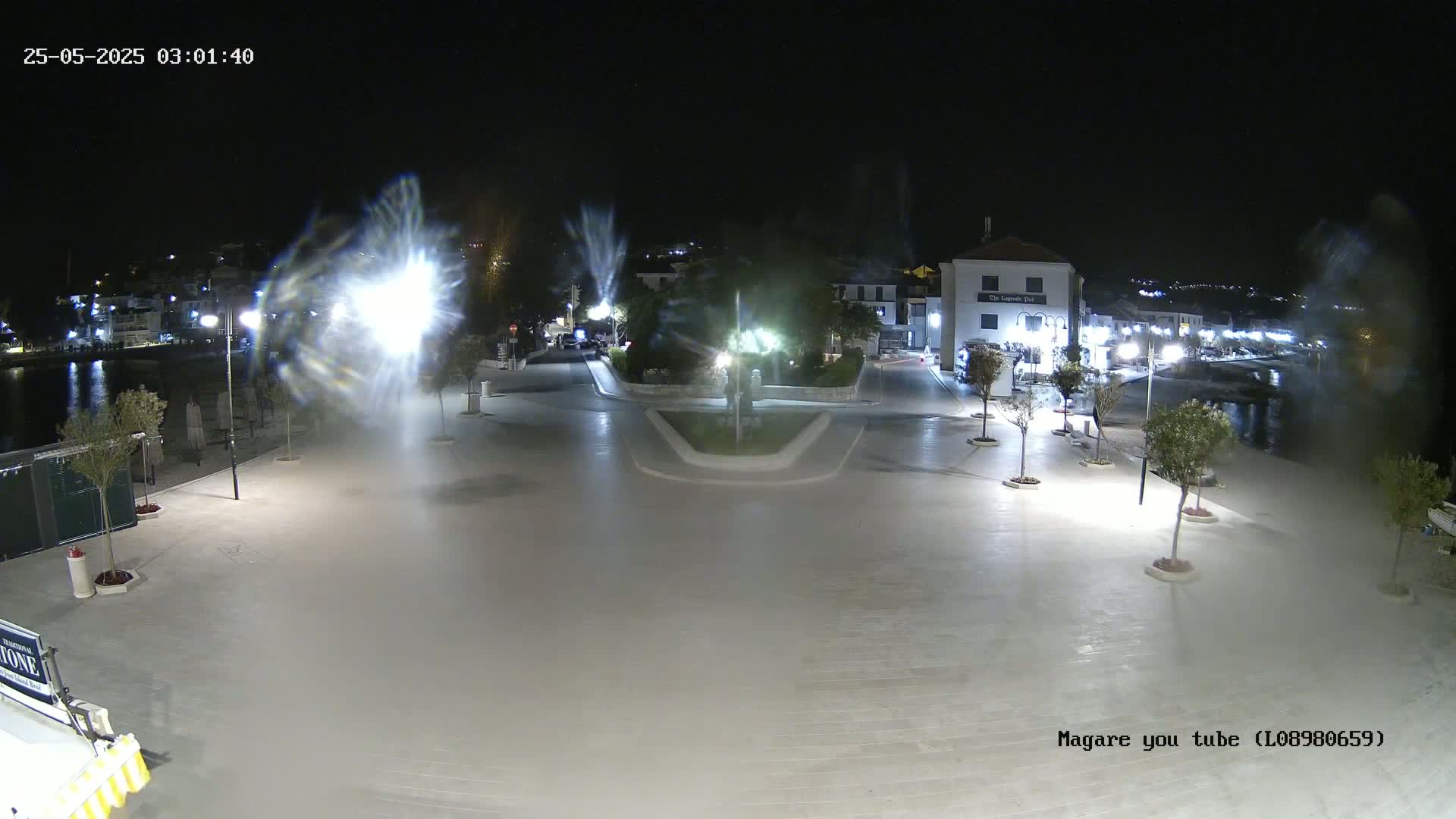 A nighttime, brightly lit town square overlooks a calm body of water, with several small trees lining the paved area.
