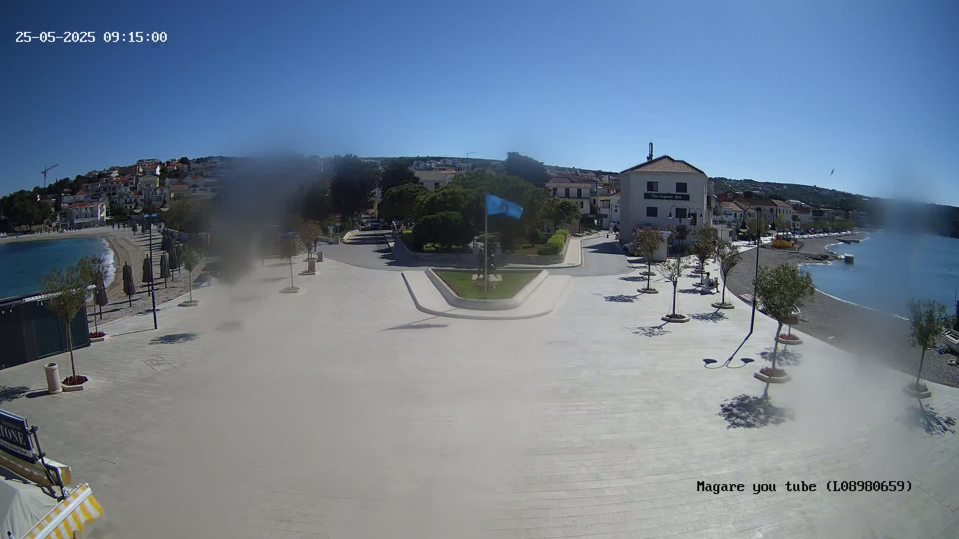 A paved seaside town square with a small park in the center,  buildings, and a calm, clear blue sea under a sunny sky.