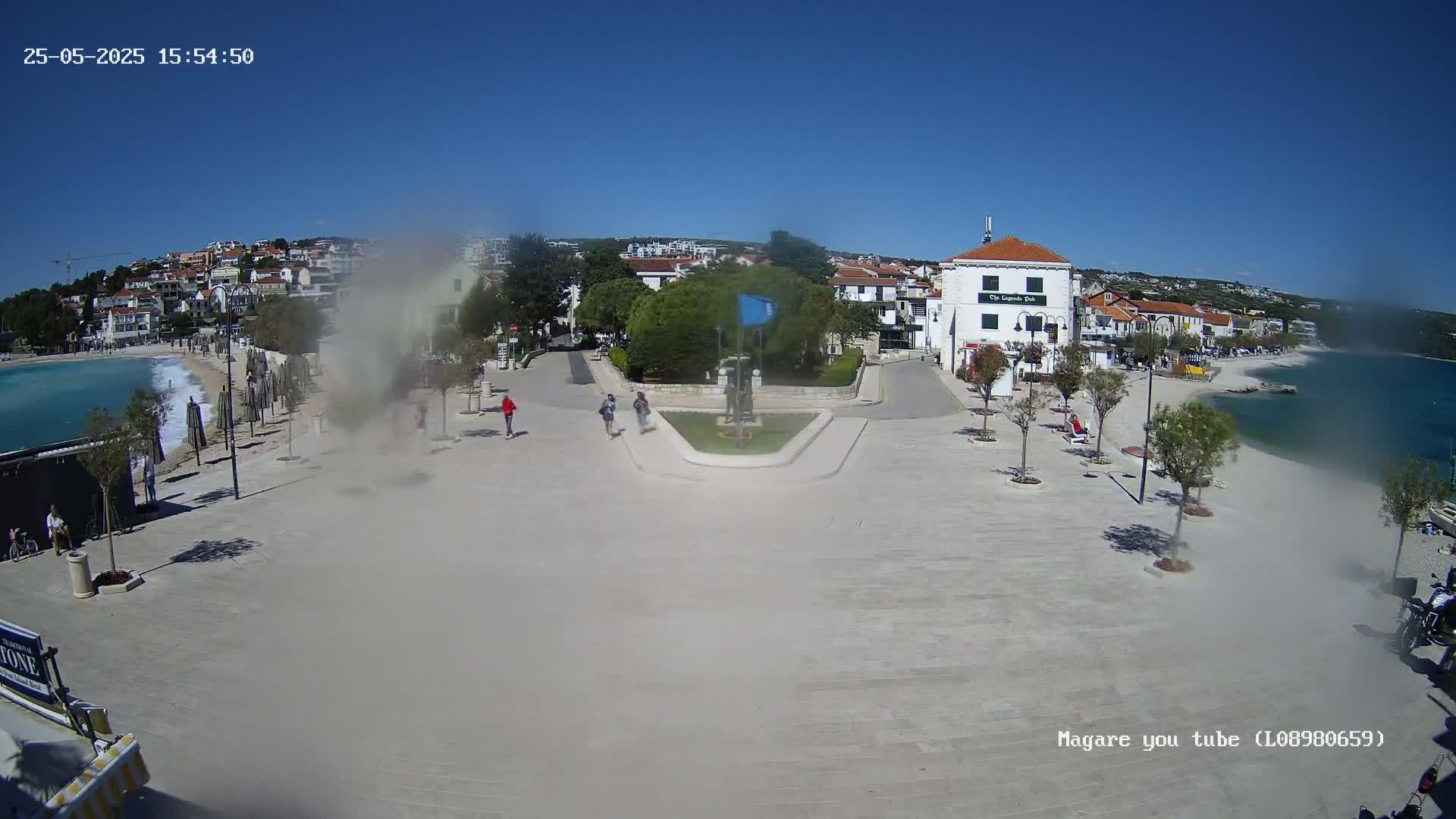 A seaside town square with a few pedestrians, small trees, and buildings, on a sunny day with some light wind blowing dust.