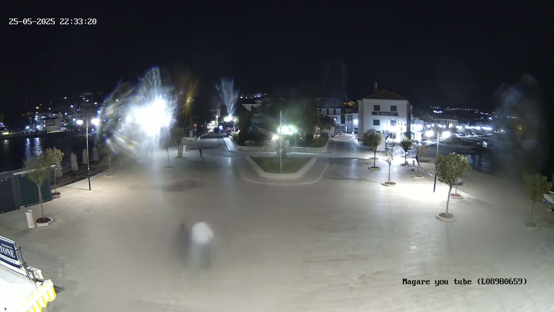 A nighttime, illuminated town square near a body of water, with a few people walking around.