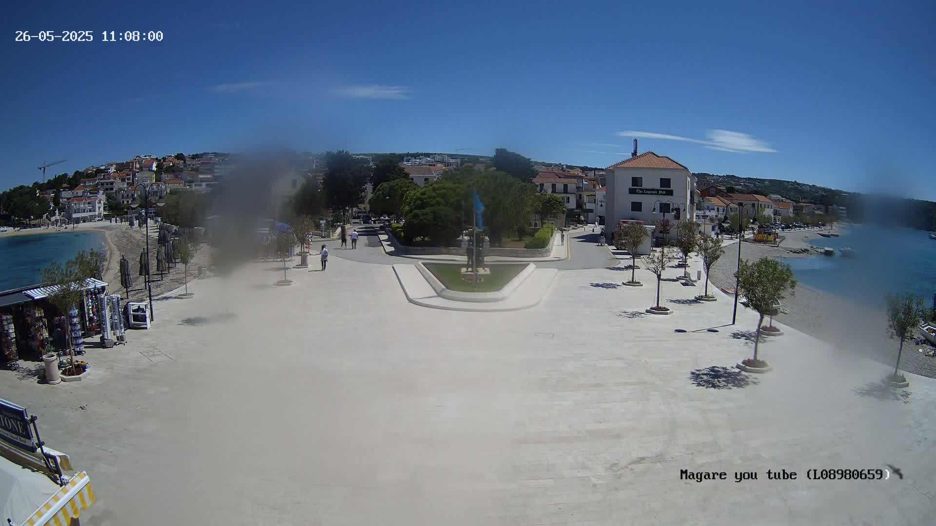 A sunny day reveals a coastal town square with a small beach, calm water, and buildings lining the shore.