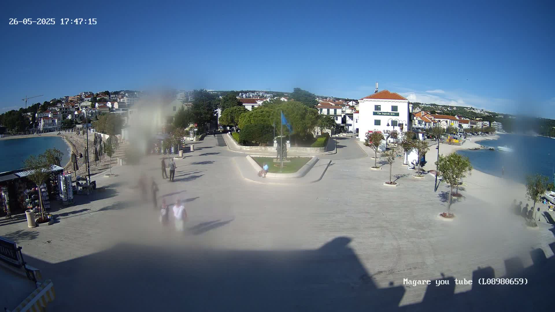 A mostly sunny seaside town square with a few people walking around, some buildings, and a small park in the center.