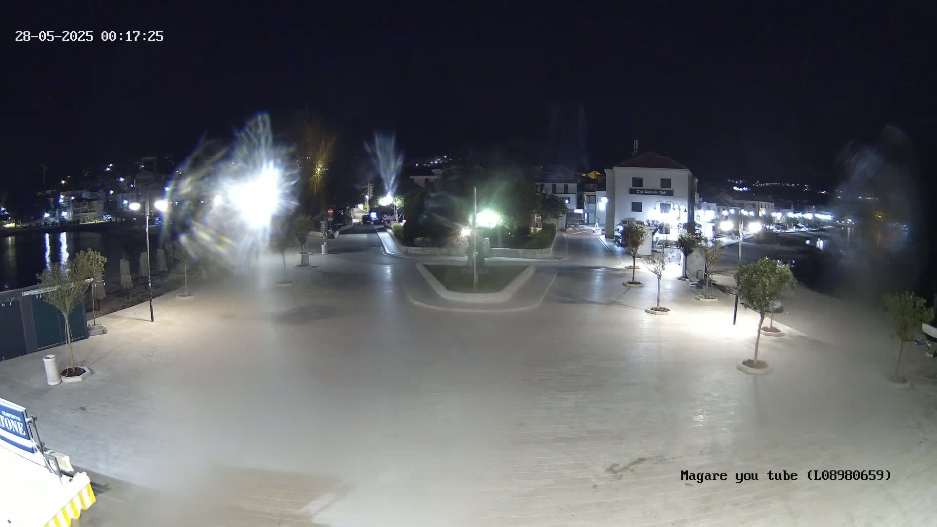 A nighttime view of a paved waterfront plaza, illuminated by streetlights, with a calm body of water in the background.