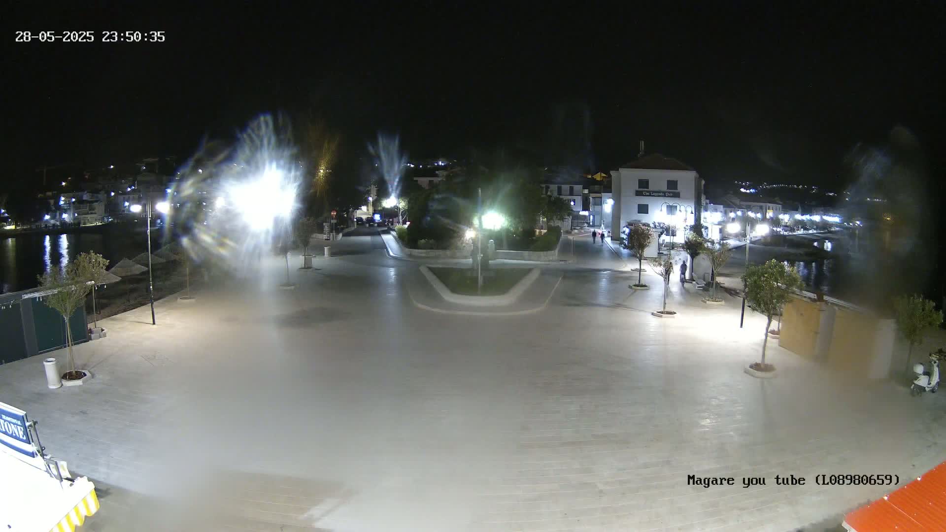 A nighttime, outdoor view of a paved town square near a body of water, with several bright lights and a mist or fog obscuring some areas.