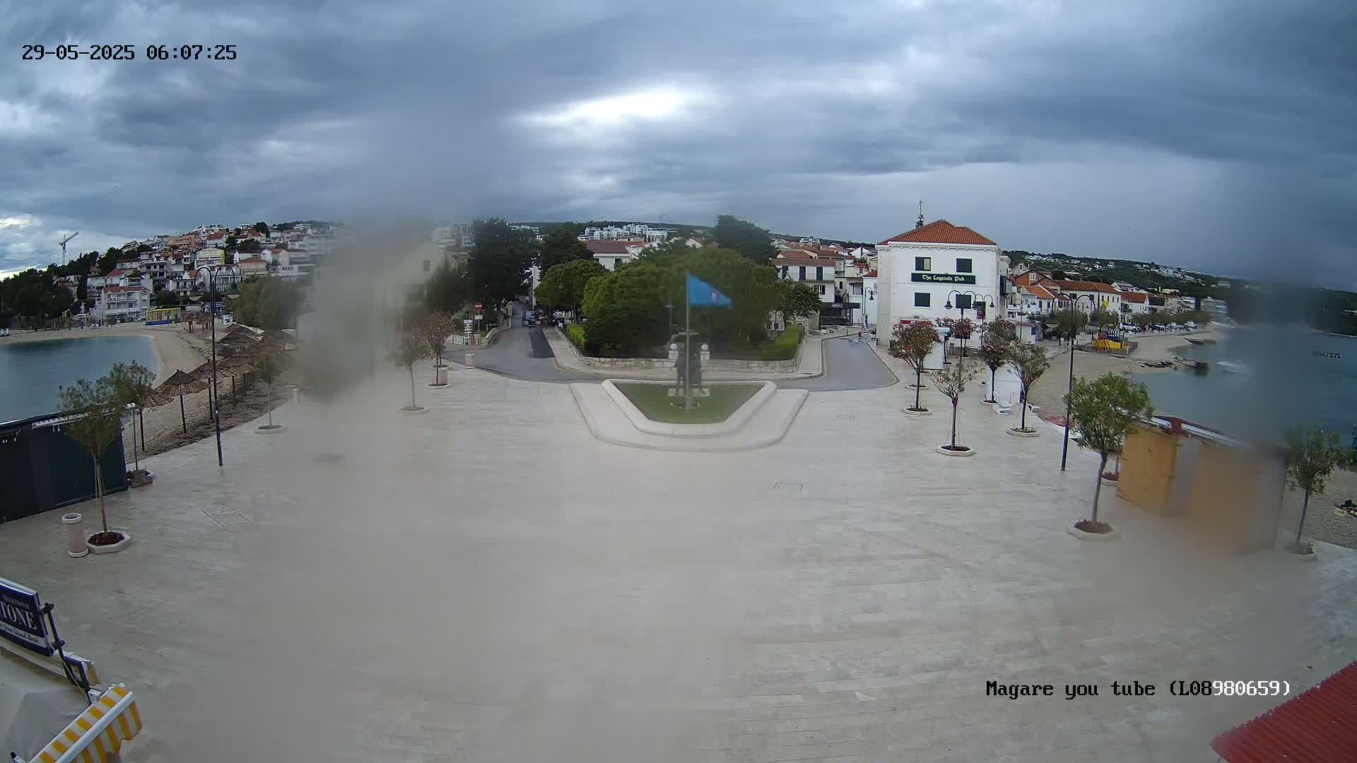 A seaside town square with overcast skies and light rain, centered around a monument and featuring buildings and a beach in the background.