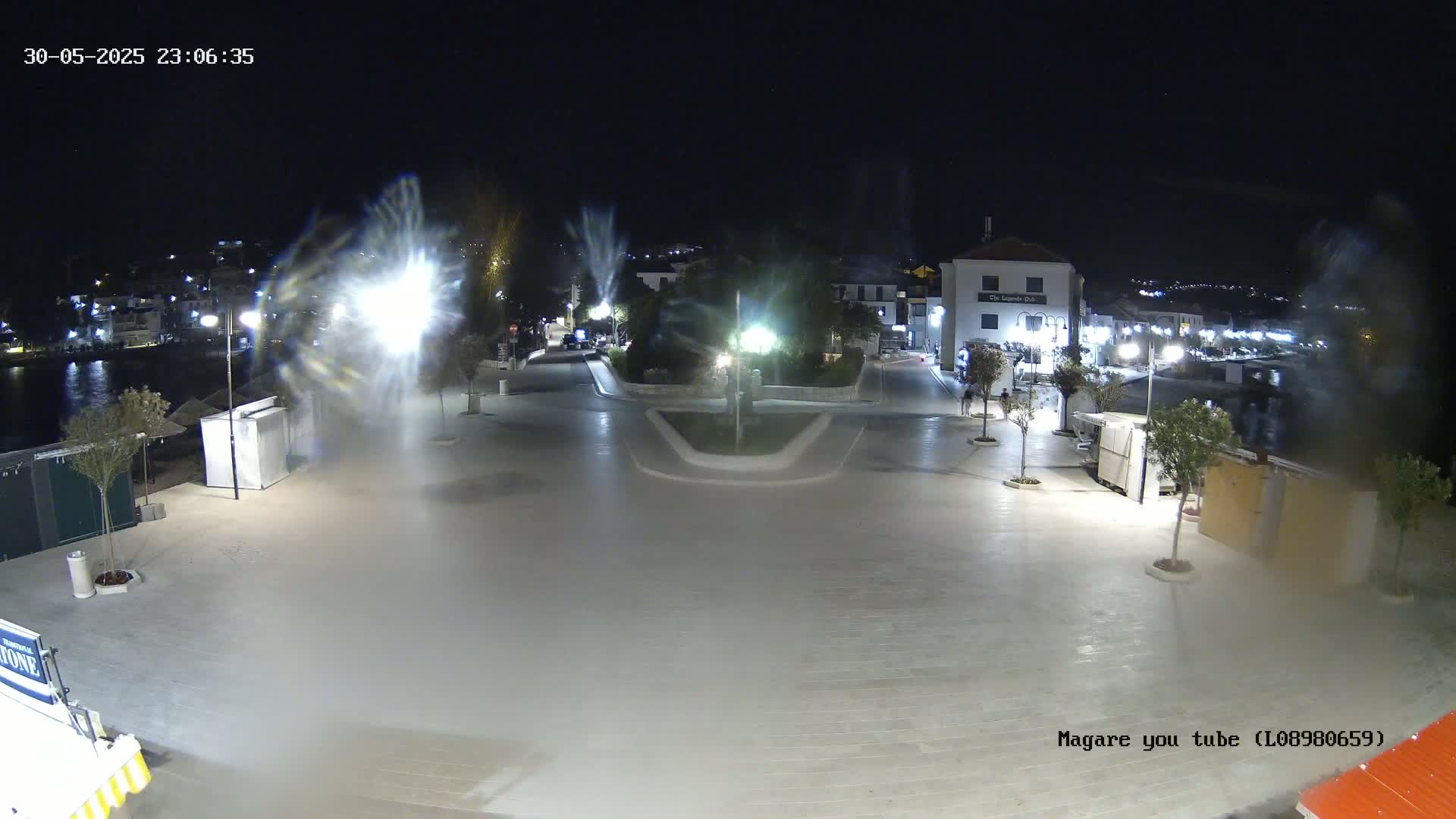 A nighttime, brightly lit plaza near a body of water, with several small trees and buildings visible in the background.