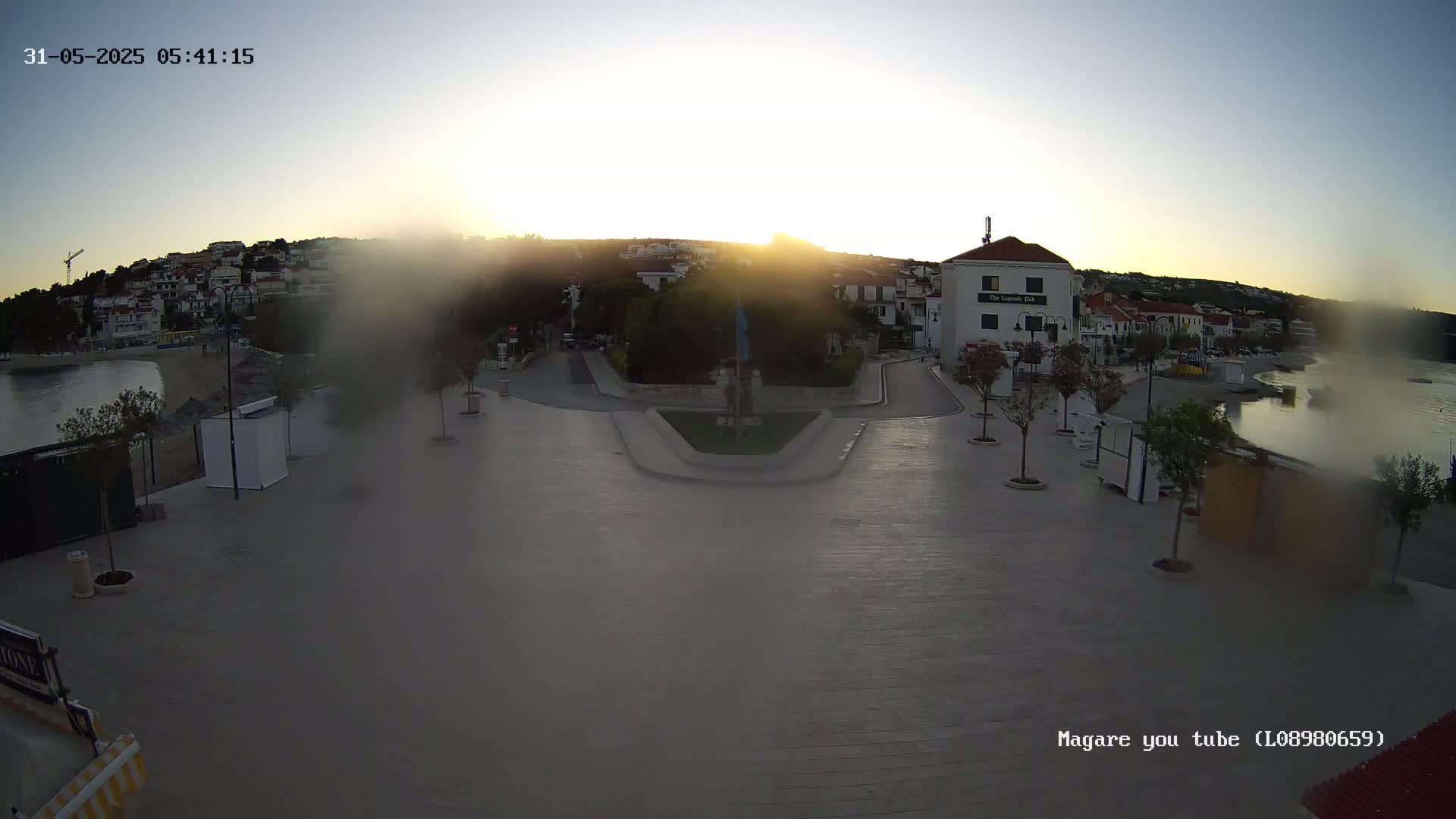 A hazy sunrise illuminates a paved town square overlooking a calm bay and a hillside of buildings.