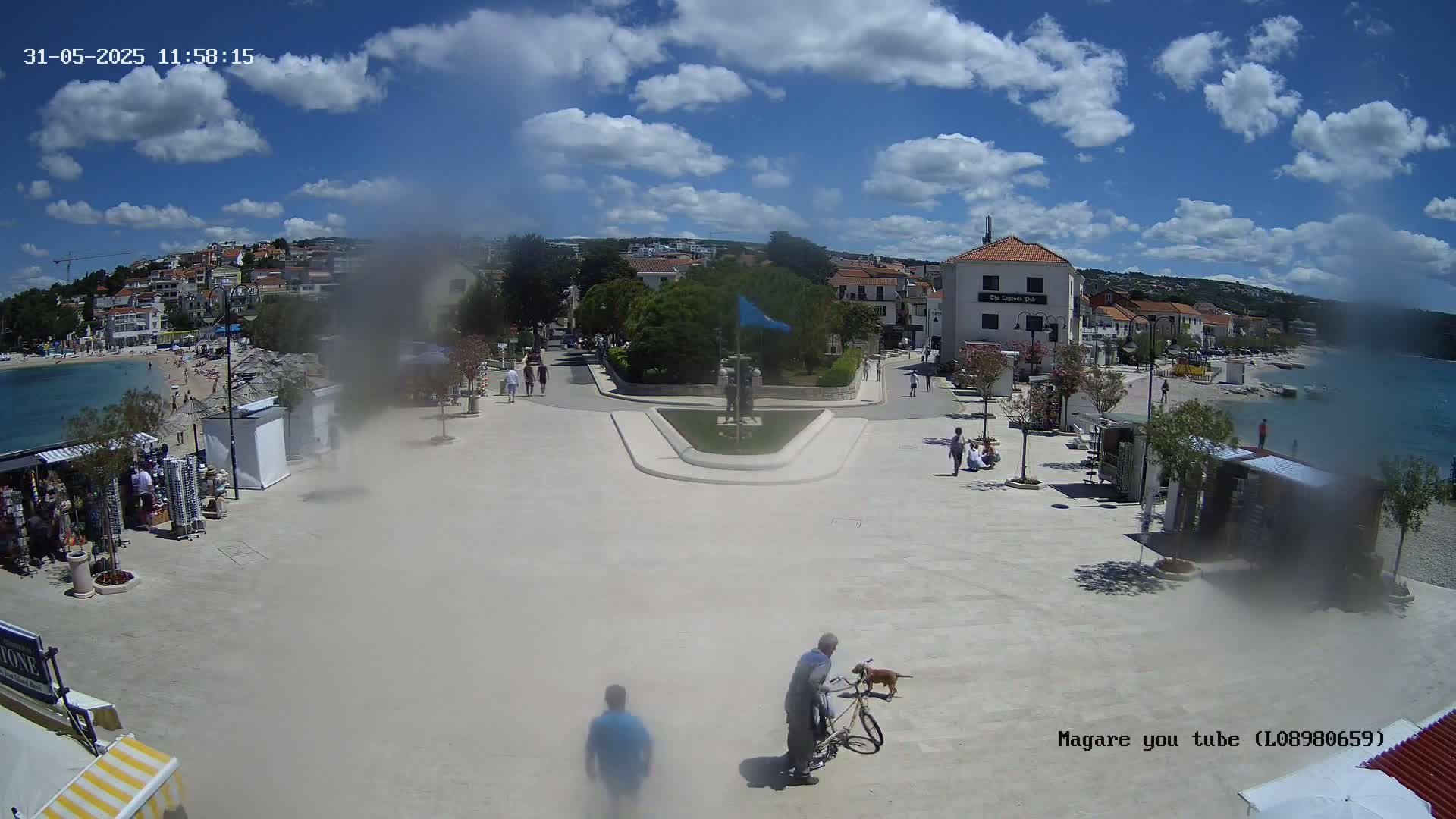 A seaside town square on a sunny day with partly cloudy skies, showing people walking around, shops, and a beach in the background.