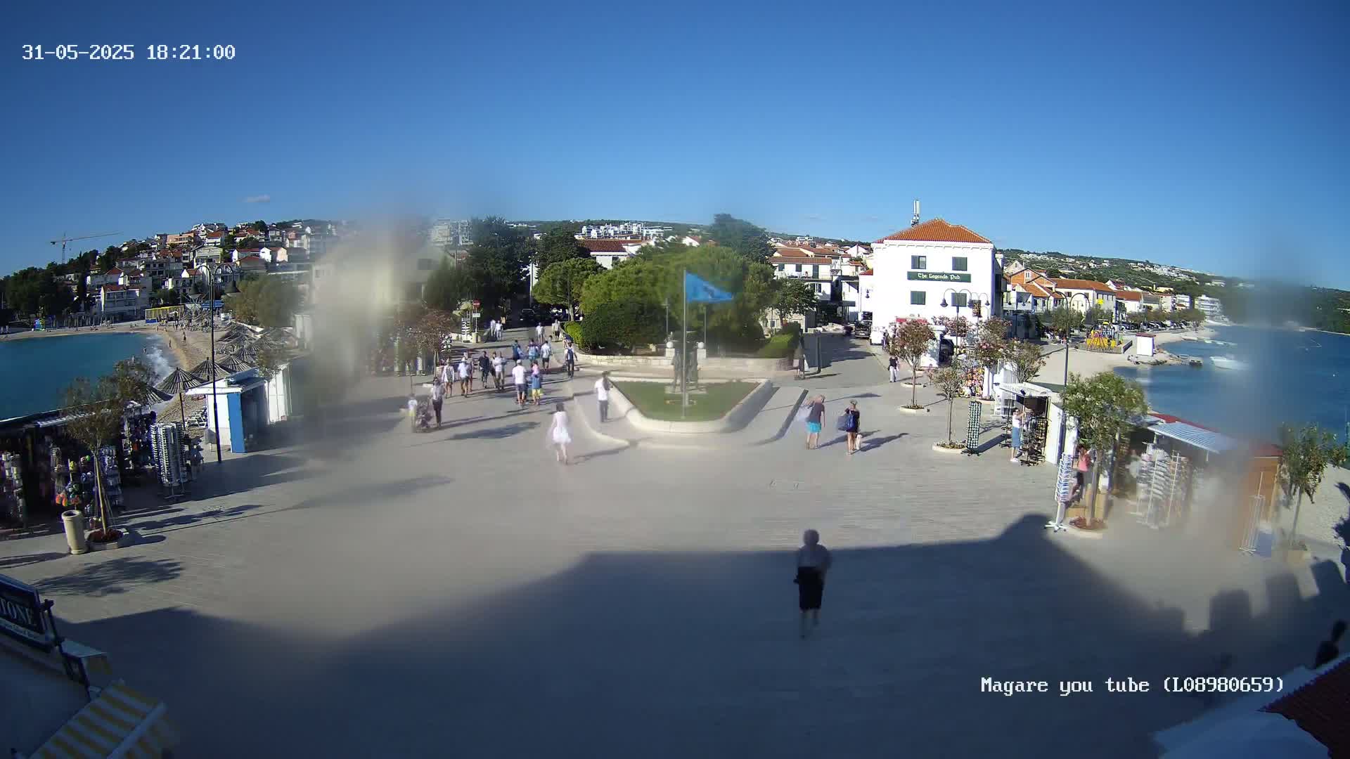 A sunny seaside town square with people walking around, shops, and a small beach visible in the background.