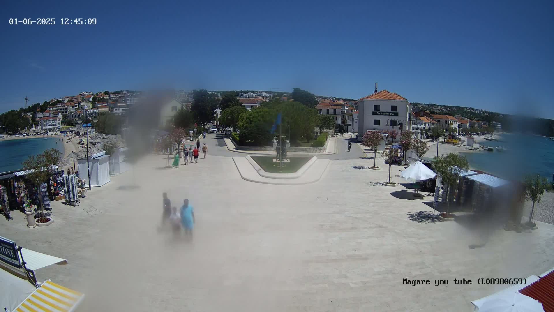 A seaside town square on a sunny day, with people walking near souvenir stands and a calm body of water in the background.