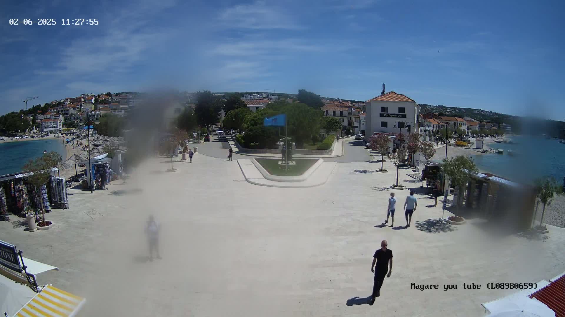 A sunny day shows a seaside town square with people walking, souvenir stands, and a view of the calm ocean.