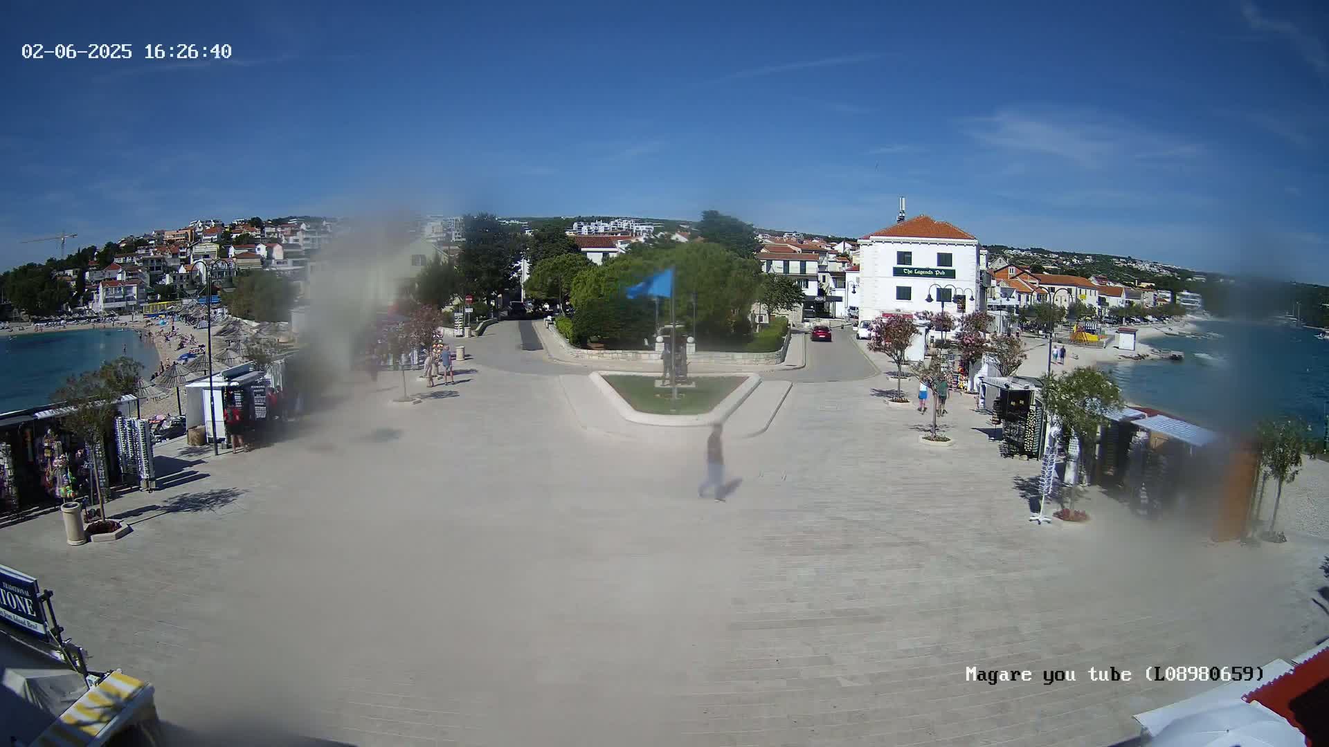 A paved town square overlooks a beach and calm ocean under a clear blue sky, with several small shops and a few people present.
