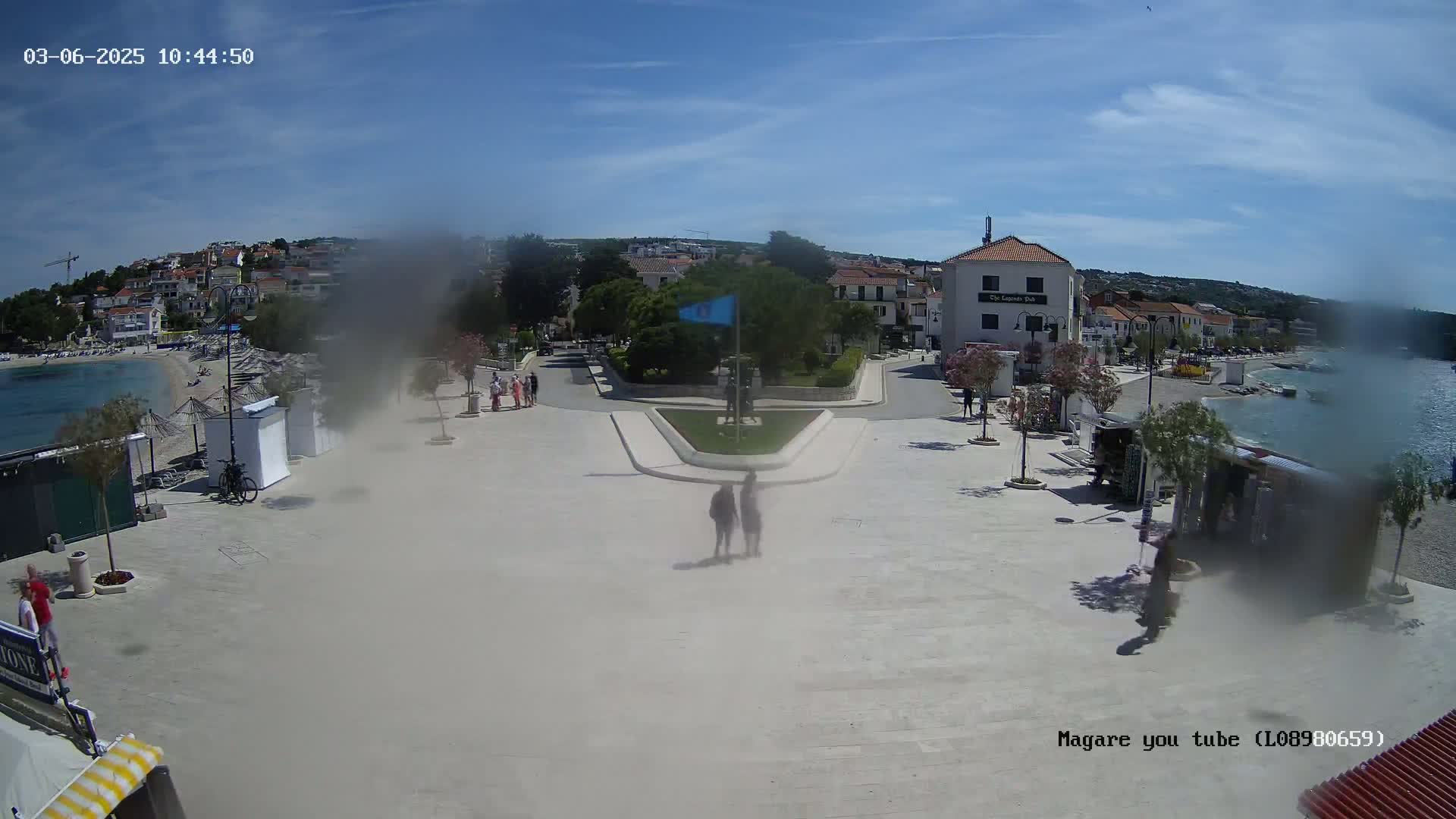 A seaside town square, mostly empty except for a few pedestrians, on a sunny day with a hazy or dusty atmosphere.