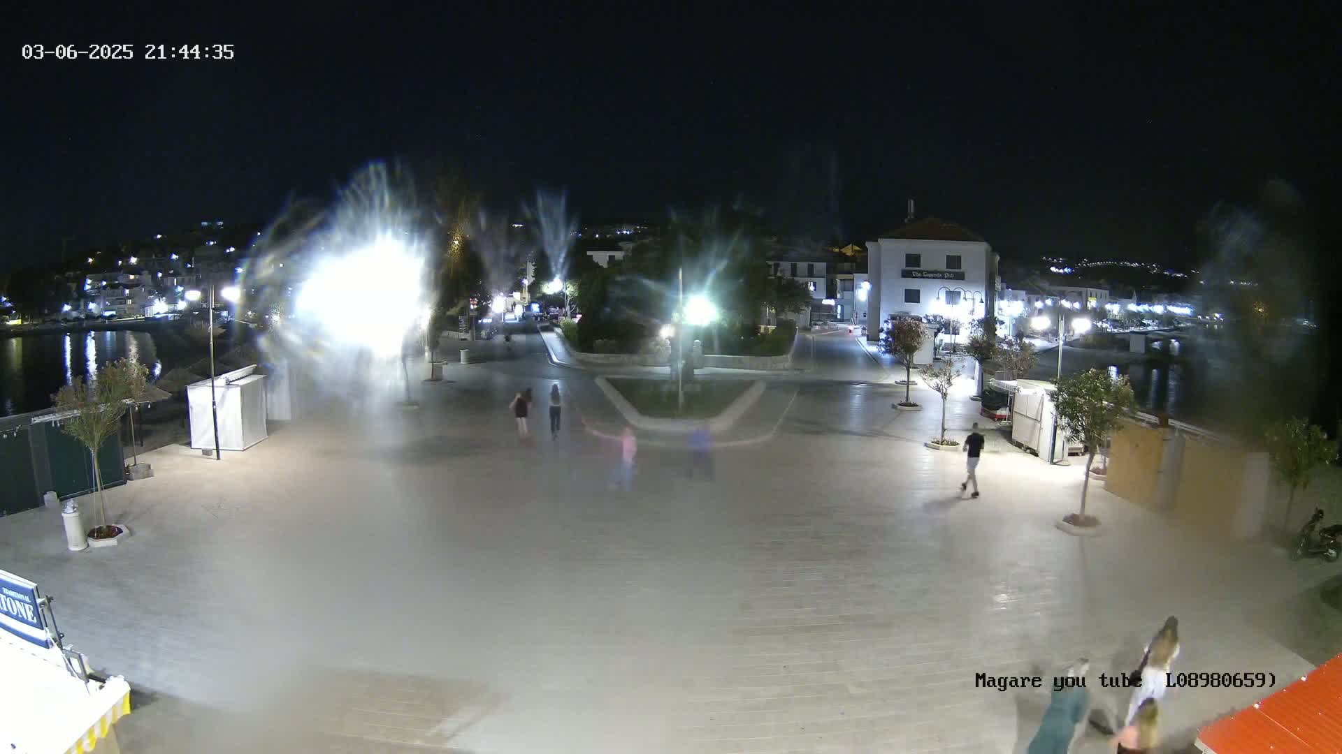 A nighttime view of a town square near a body of water, with several people walking around and bright lights creating a hazy effect in the air.