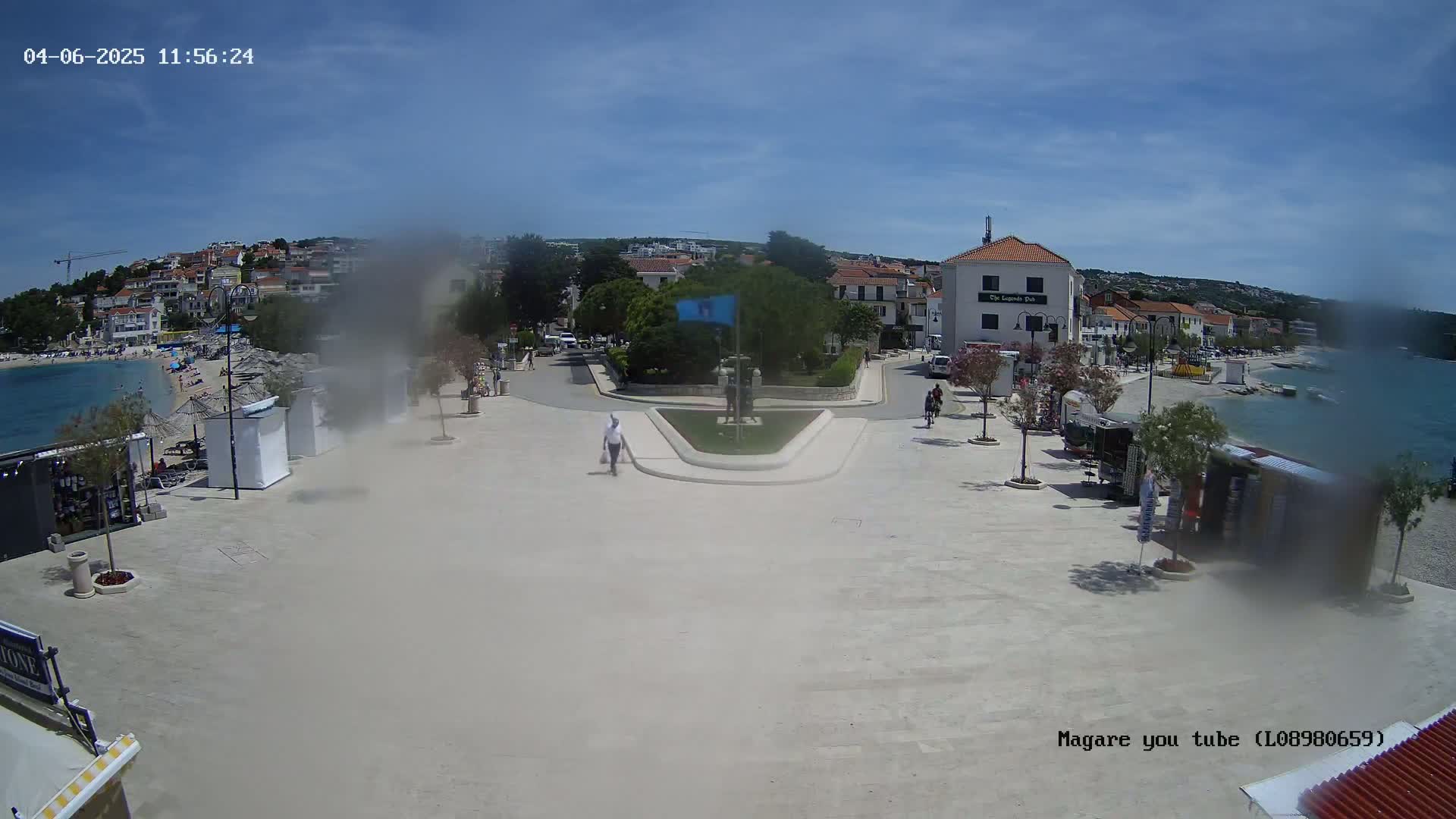 A paved plaza overlooks a calm bay on a sunny day, with buildings lining the shore and small kiosks scattered around the plaza.