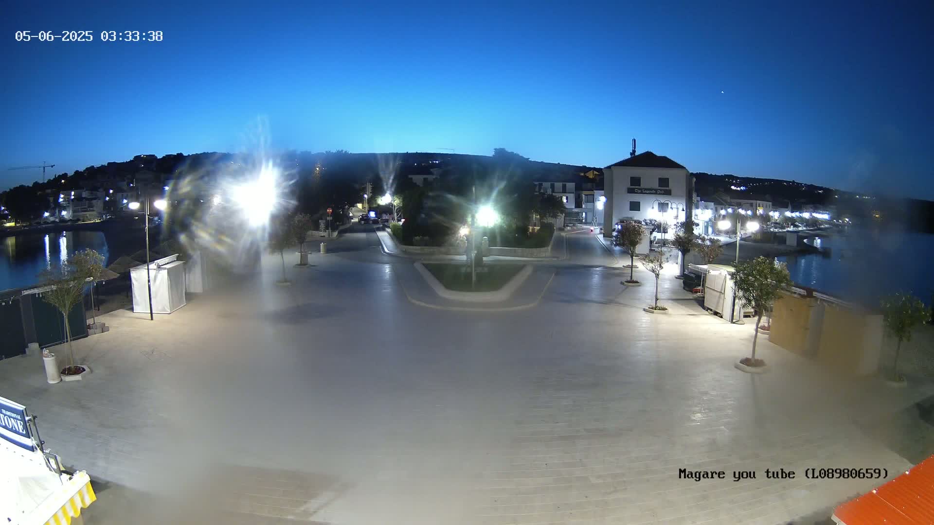 A nighttime, slightly hazy view of an empty town square near a calm body of water, with buildings and trees lining the perimeter under bright artificial lights.