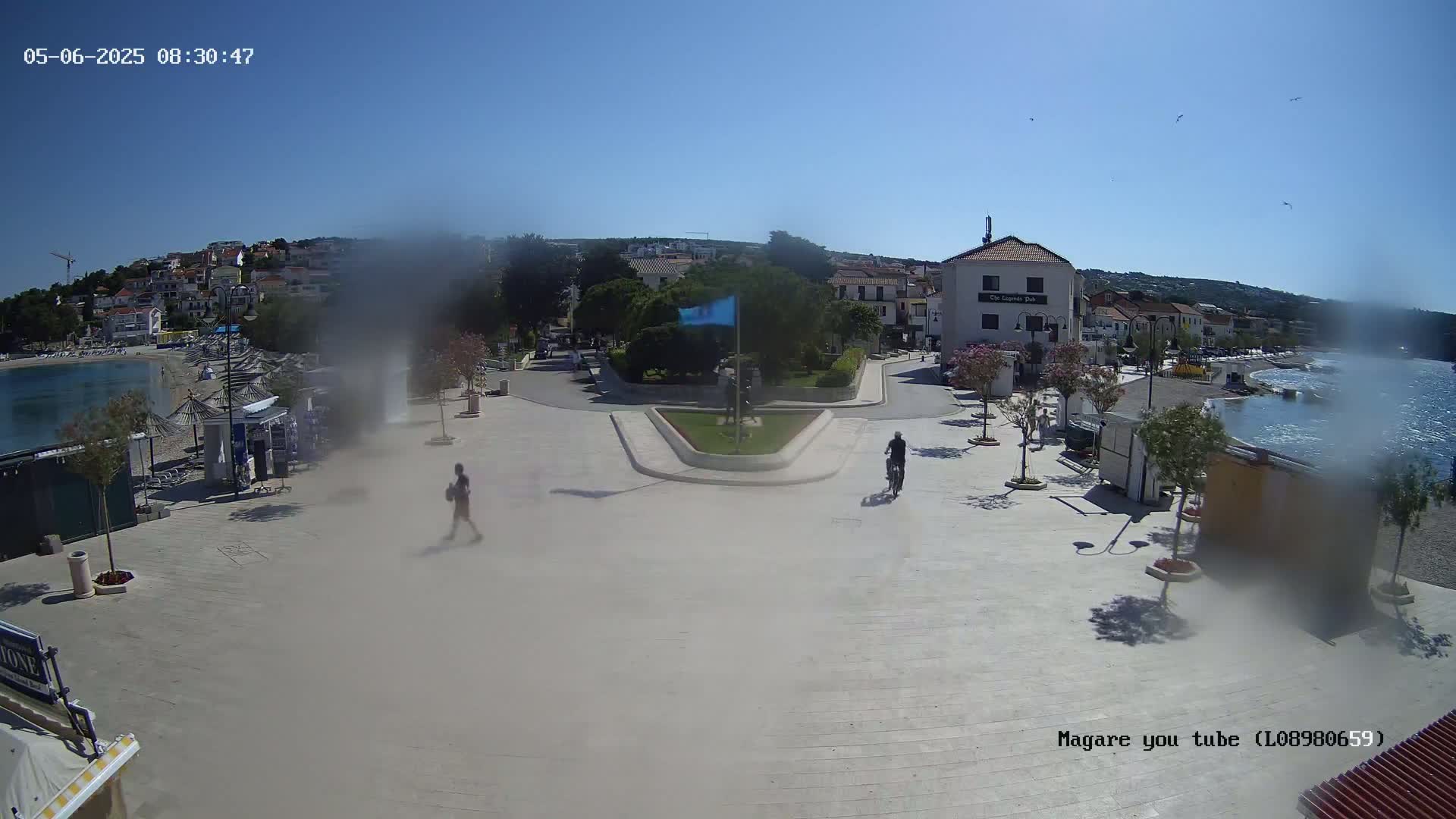 A hazy, sunny day shows a paved town square near a calm body of water, with buildings, trees, and a few people visible.