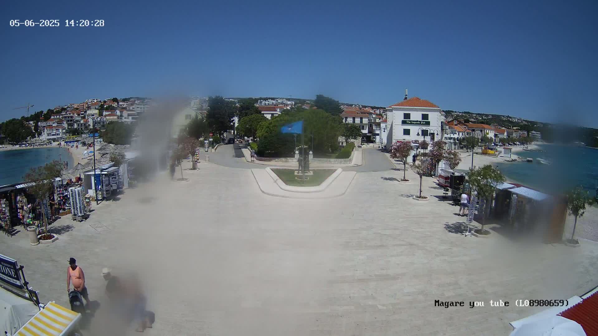 A paved town square with souvenir stands and buildings bordering a calm, blue body of water under a clear, sunny sky.
