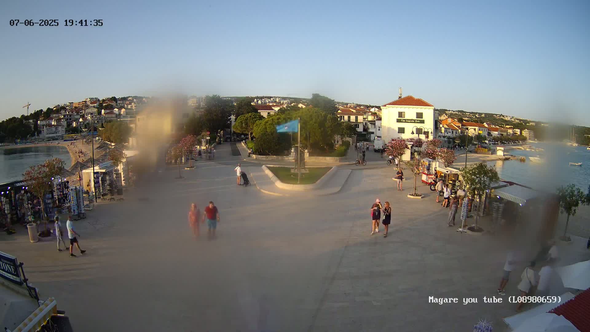 A hazy, sunlit seaside town square with several people walking past souvenir stands near a calm body of water, under a clear sky.