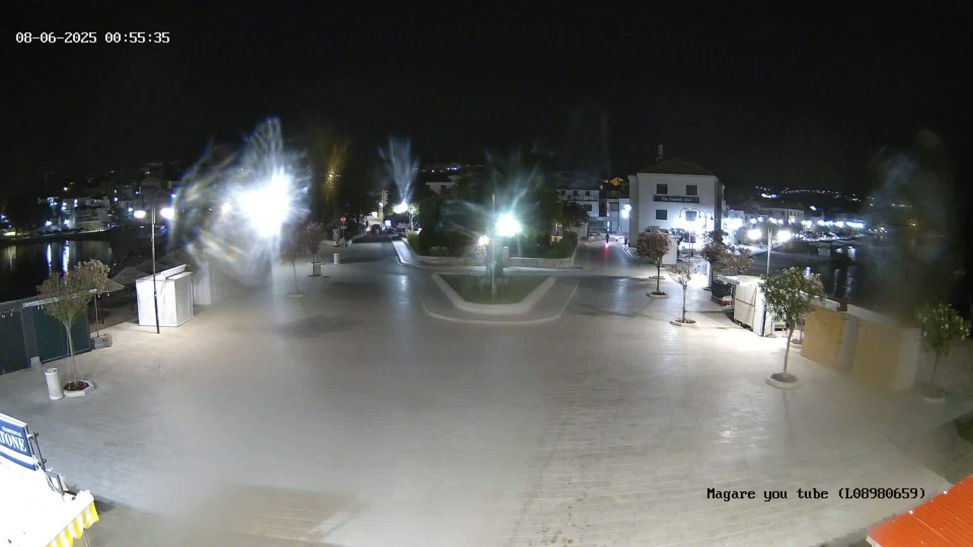 A nighttime view of a town square near a body of water, illuminated by streetlights, with some light fog or mist.