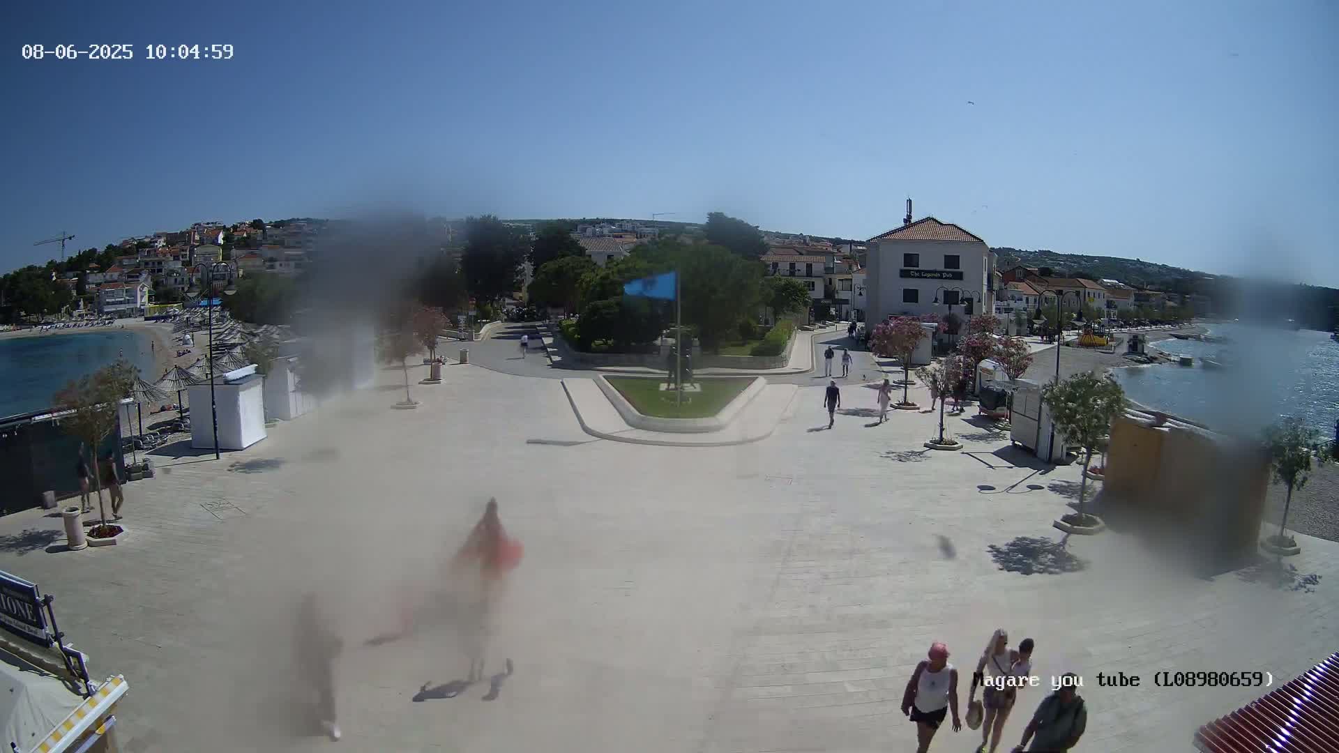 A hazy, sunny day overlooks a town square near a calm body of water with several people walking around.