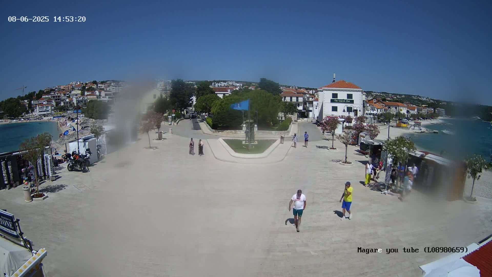 A paved plaza near a beach on a sunny day, with buildings, people walking, and some small shops or stands.