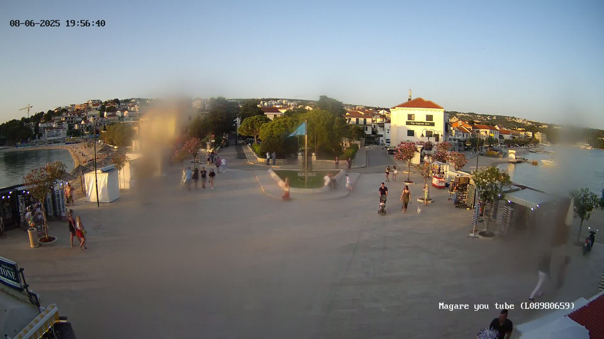 A seaside town square, under a clear evening sky, features people walking around small shops and kiosks near a calm body of water.